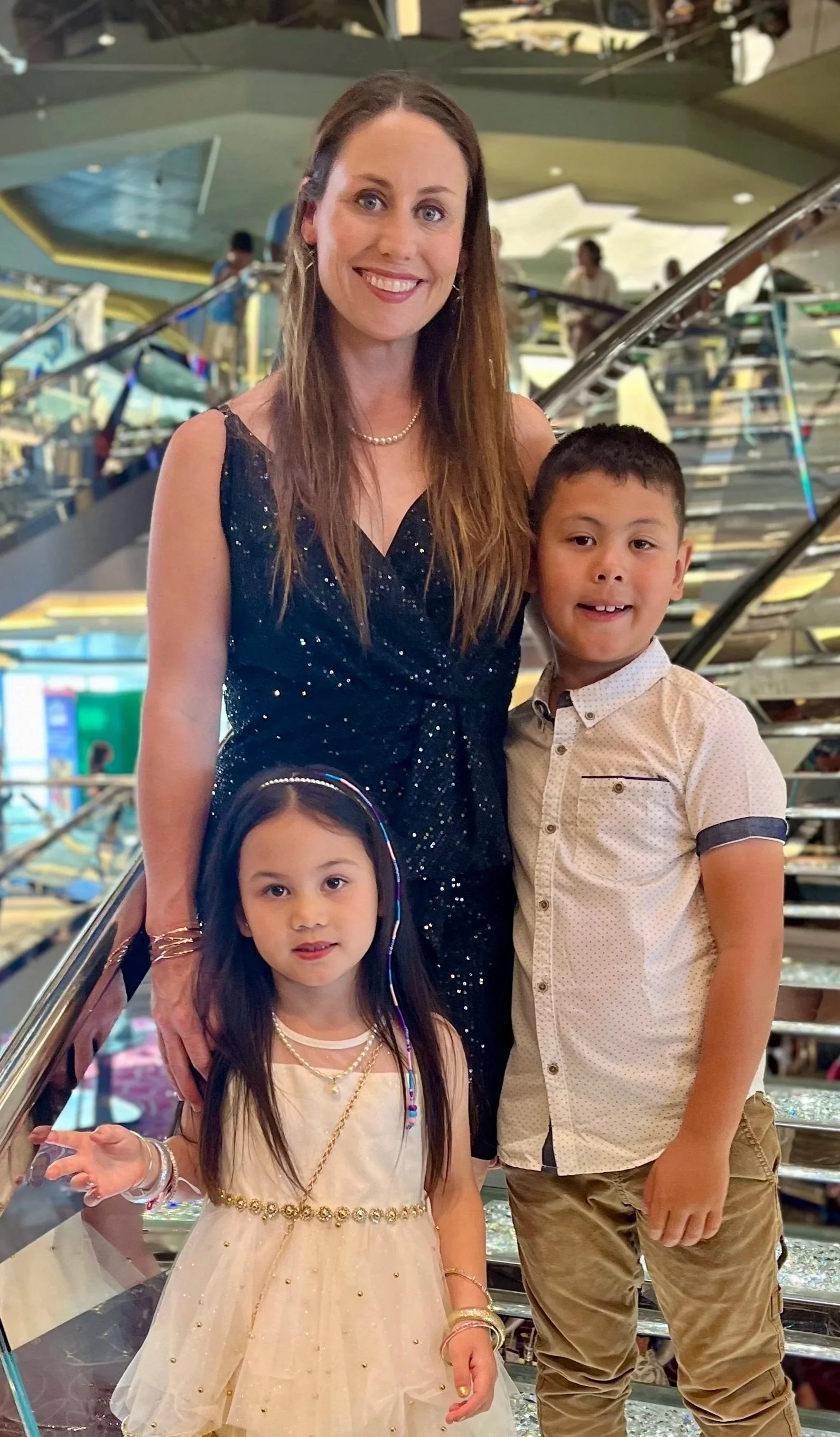 A woman in a dark blue, sparkling dress stands with three children in an indoor setting with a glass staircase and other people in the background.