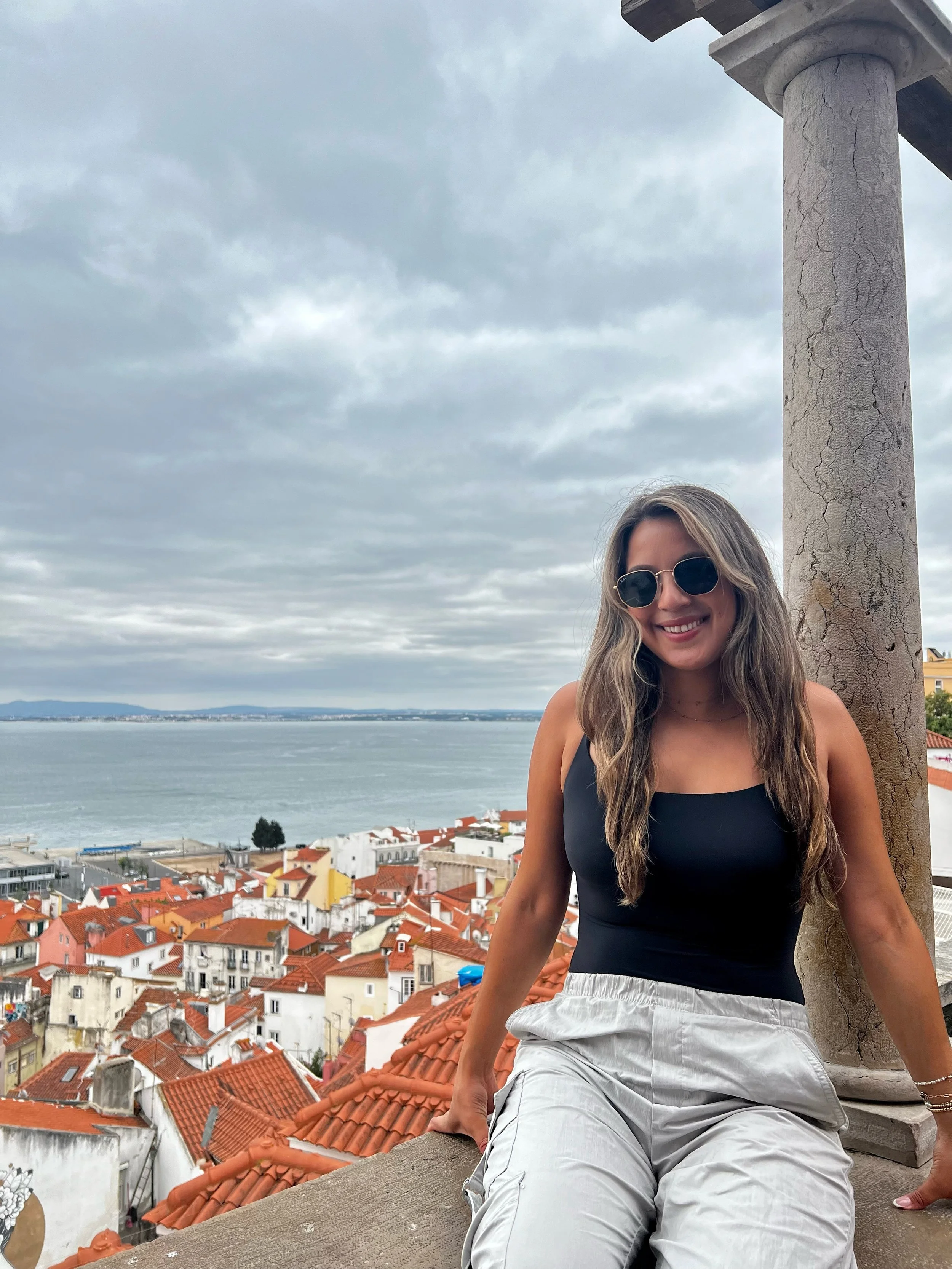 A woman with long wavy hair wearing sunglasses, a black top, and light-colored shorts, sitting on a ledge on a rooftop with a view of a city with orange rooftops, a body of water, and a cloudy sky behind her.