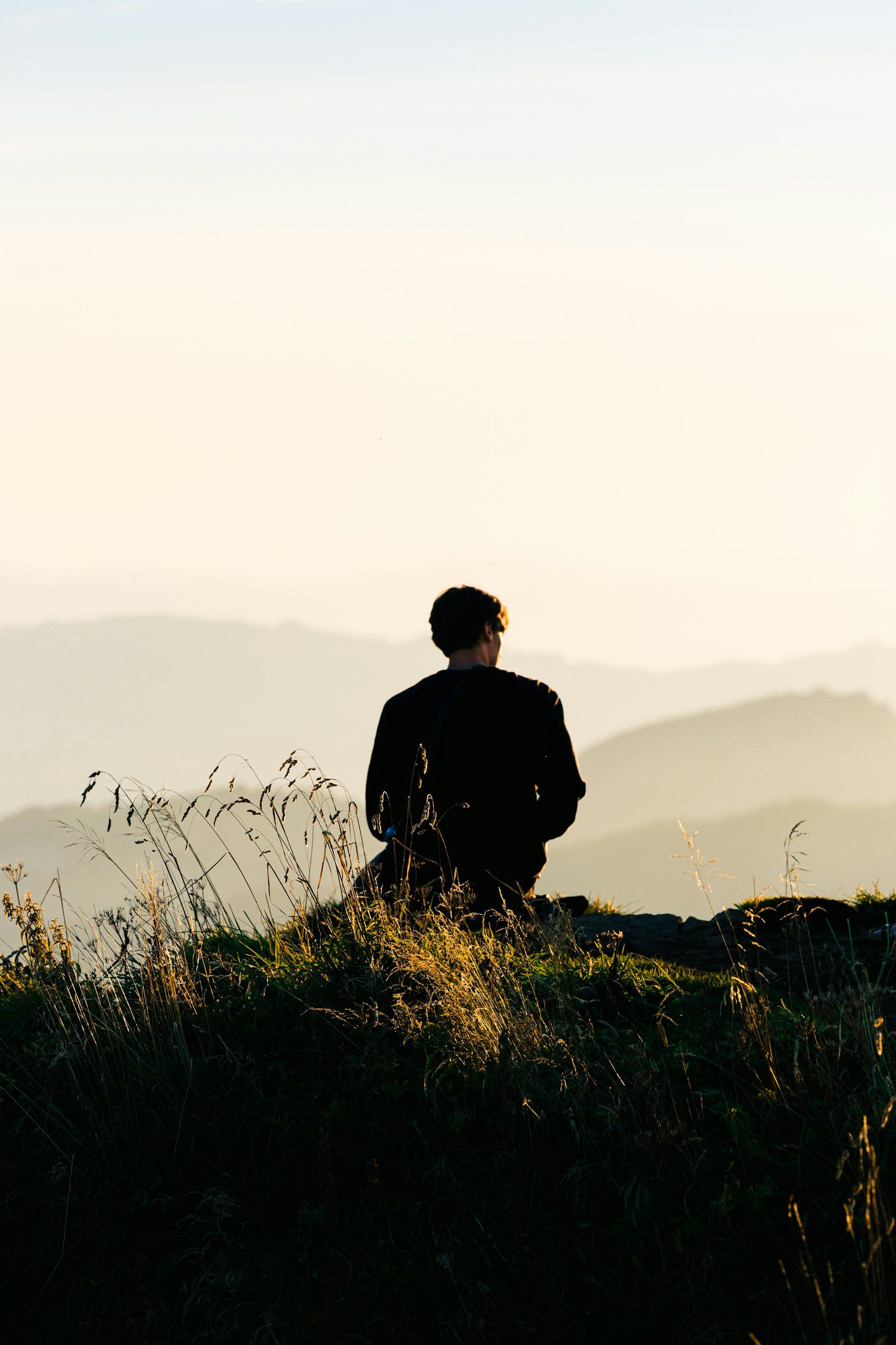 Person sitting on a hilltop during sunset, facing away, surrounded by tall grass.