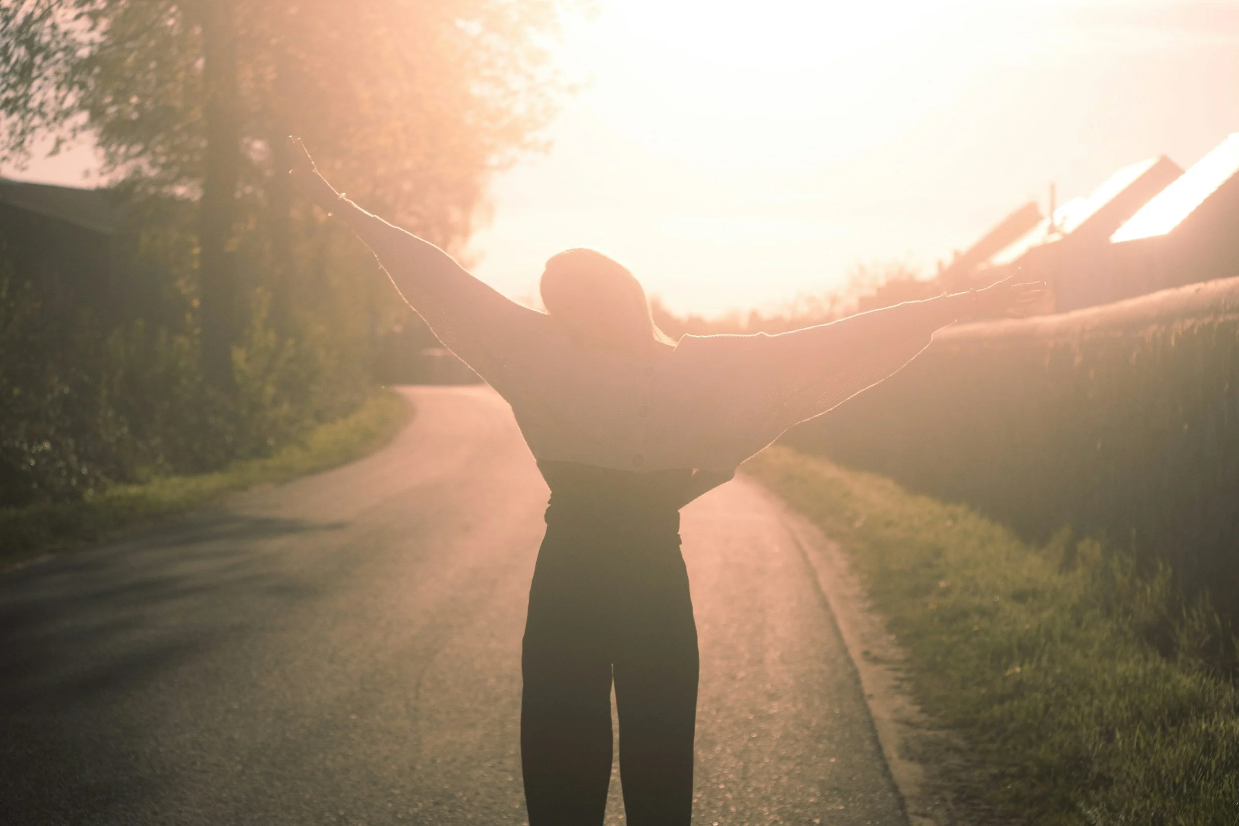 Person with arms spread wide standing on a road during sunset with trees and houses in the background.