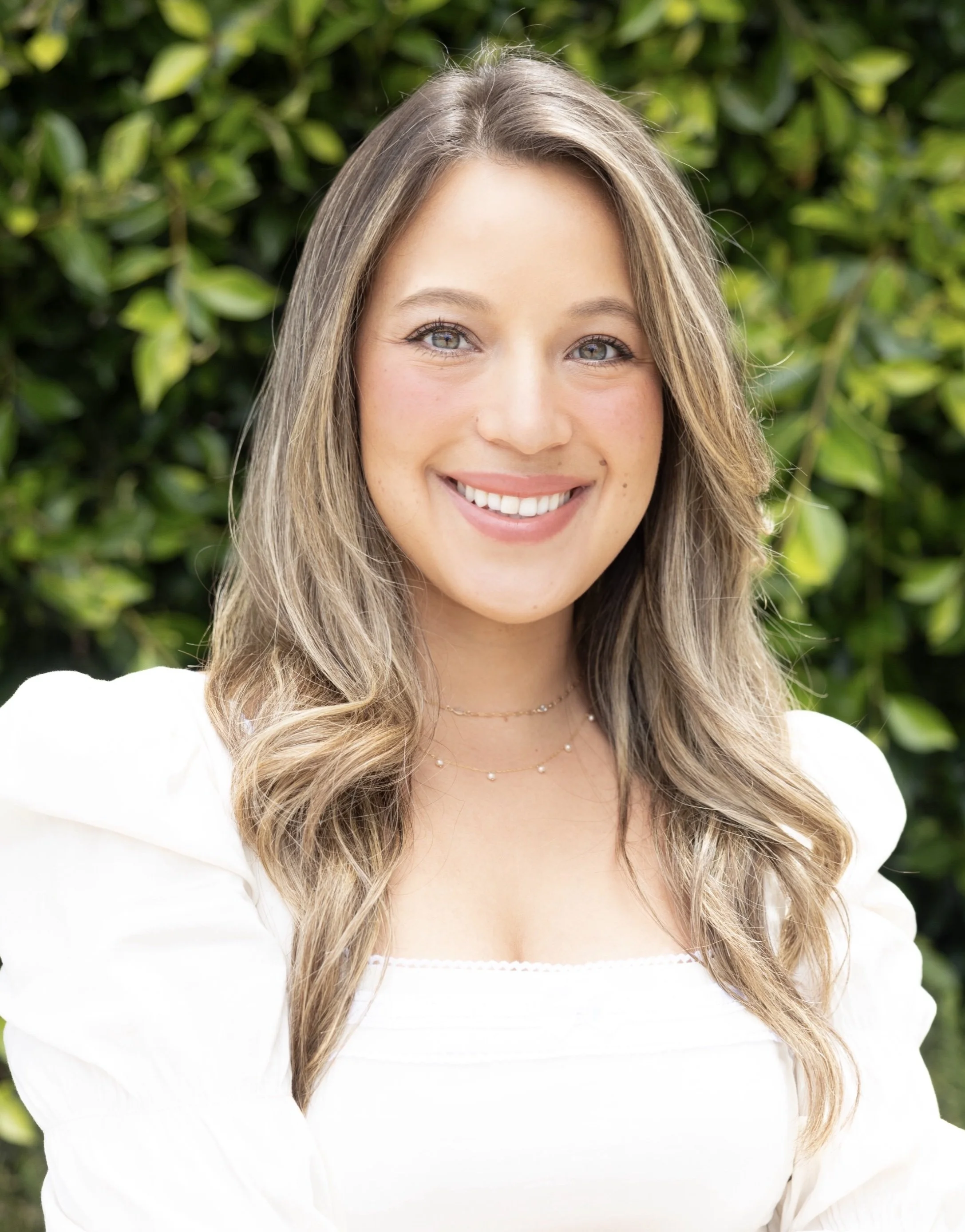 A young woman with long, wavy blonde hair, smiling, wearing a white top and jewelry, standing outdoors with green leafy background.