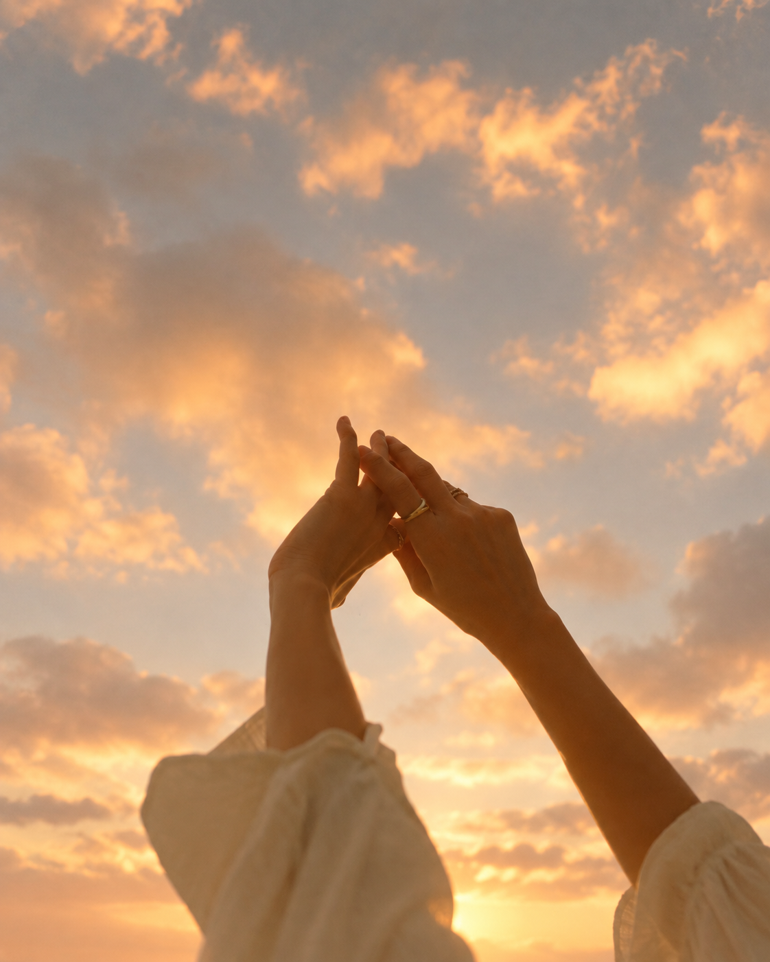 A person's hands forming a heart shape against a sunset sky with orange and pink clouds.