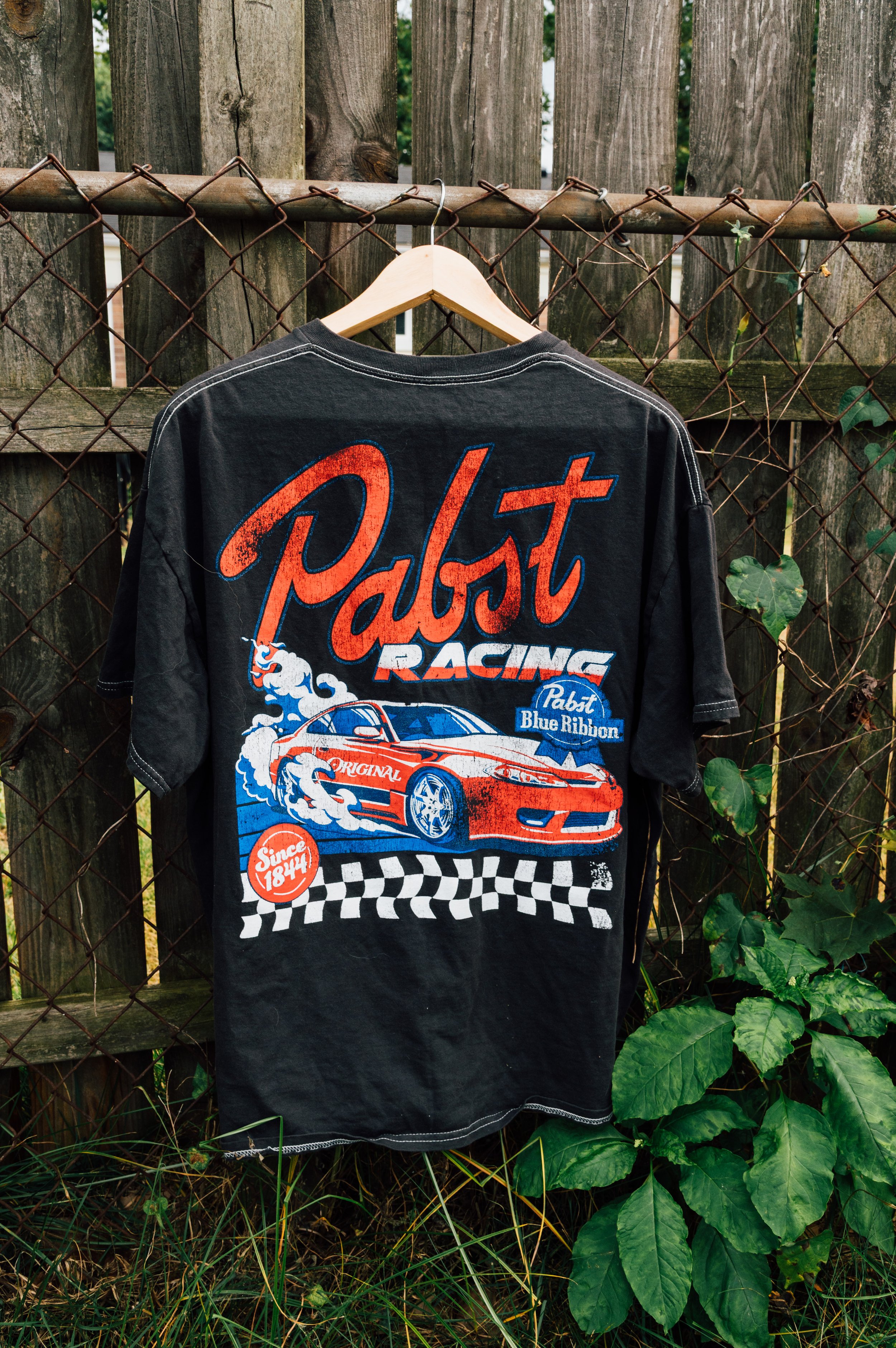 Black racing T-shirt with a graphic of a red sports car, checkered flag pattern, and text, hanging on a hanger in front of a wooden fence and green foliage.