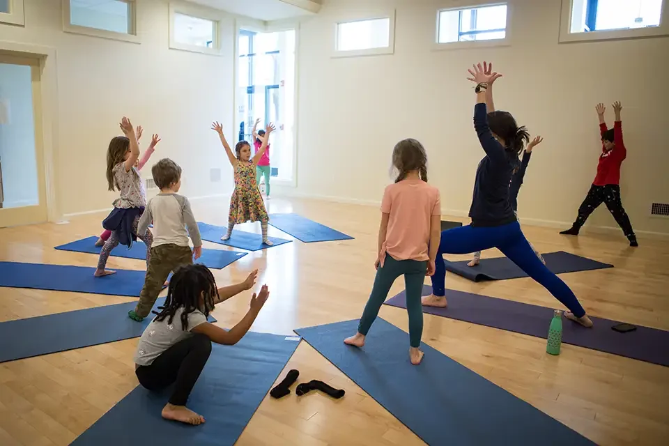 Children participating in a yoga class, stretching on mats in a bright room with large windows.