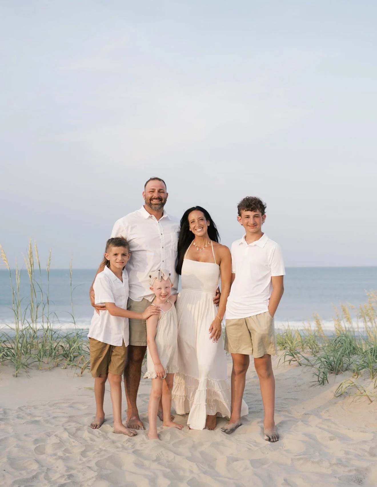 A family of six standing on a sandy beach with the ocean and sky in the background. The dad, mom, three children, and a baby girl are all smiling and dressed in light-colored, casual clothing suitable for warm weather.