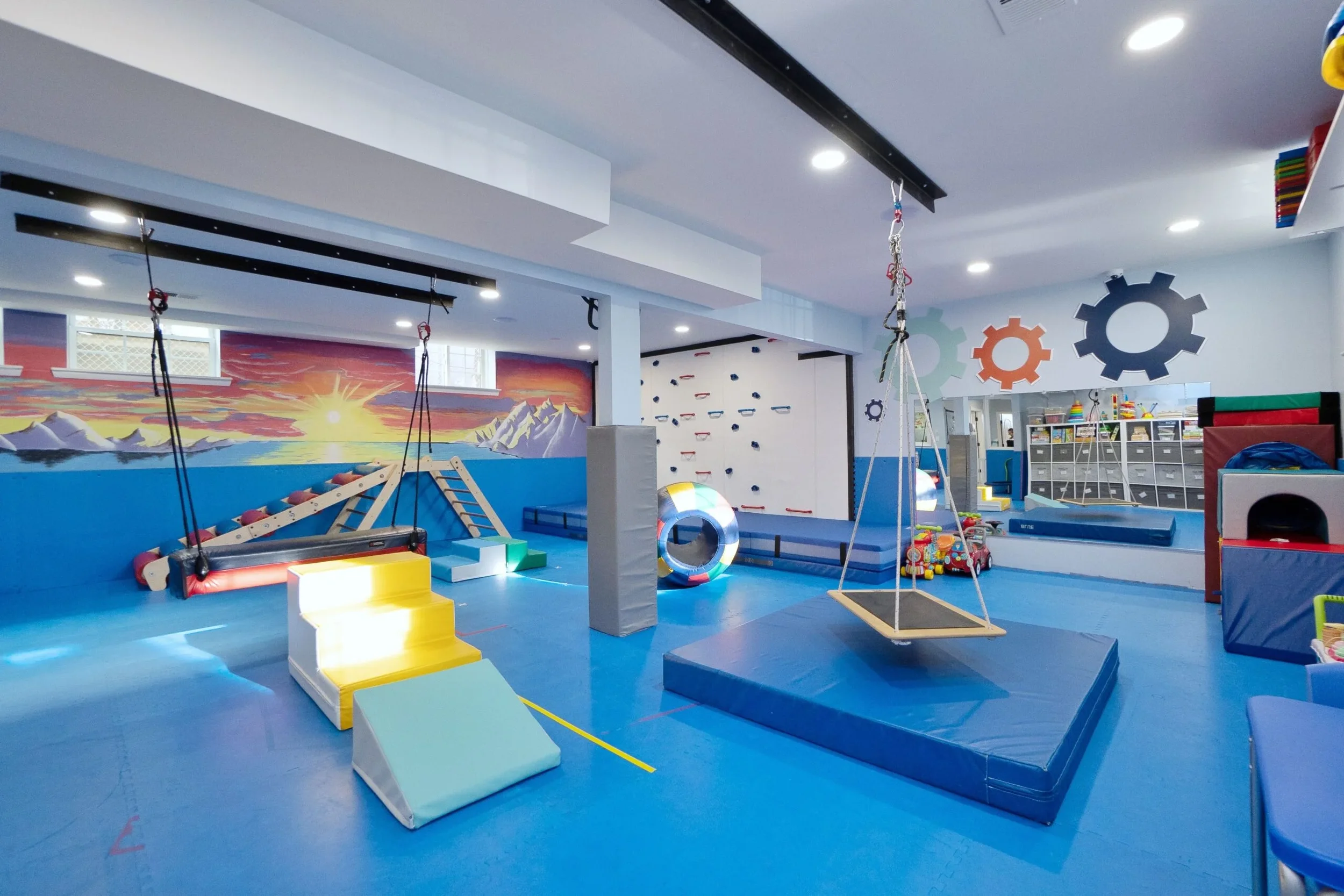 Colorful indoor children's play area with climbing wall, swings, padded mats, and various toys, featuring a mural of a sunset over mountains.