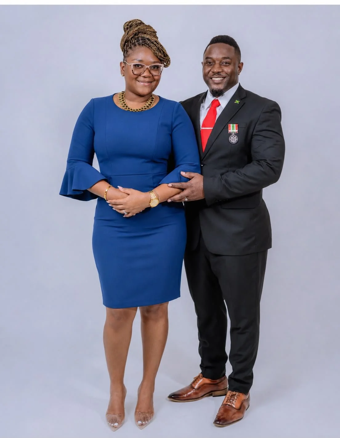 A man and woman dressed in formal attire standing together against a plain gray background. The woman is wearing a blue dress with bell sleeves, glasses, and jewelry, while the man is wearing a black suit with a red tie and medals on his chest.