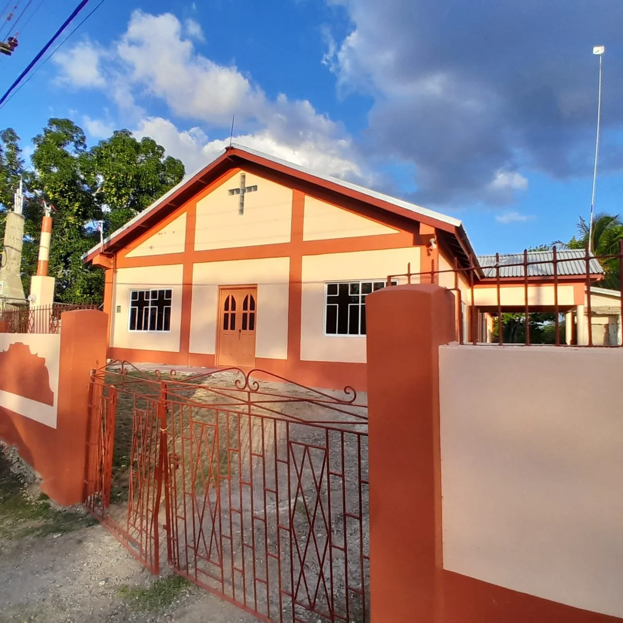 A small church with a cross on the front gable, orange and beige exterior, surrounded by a metal gate, with a bright sky and clouds overhead.