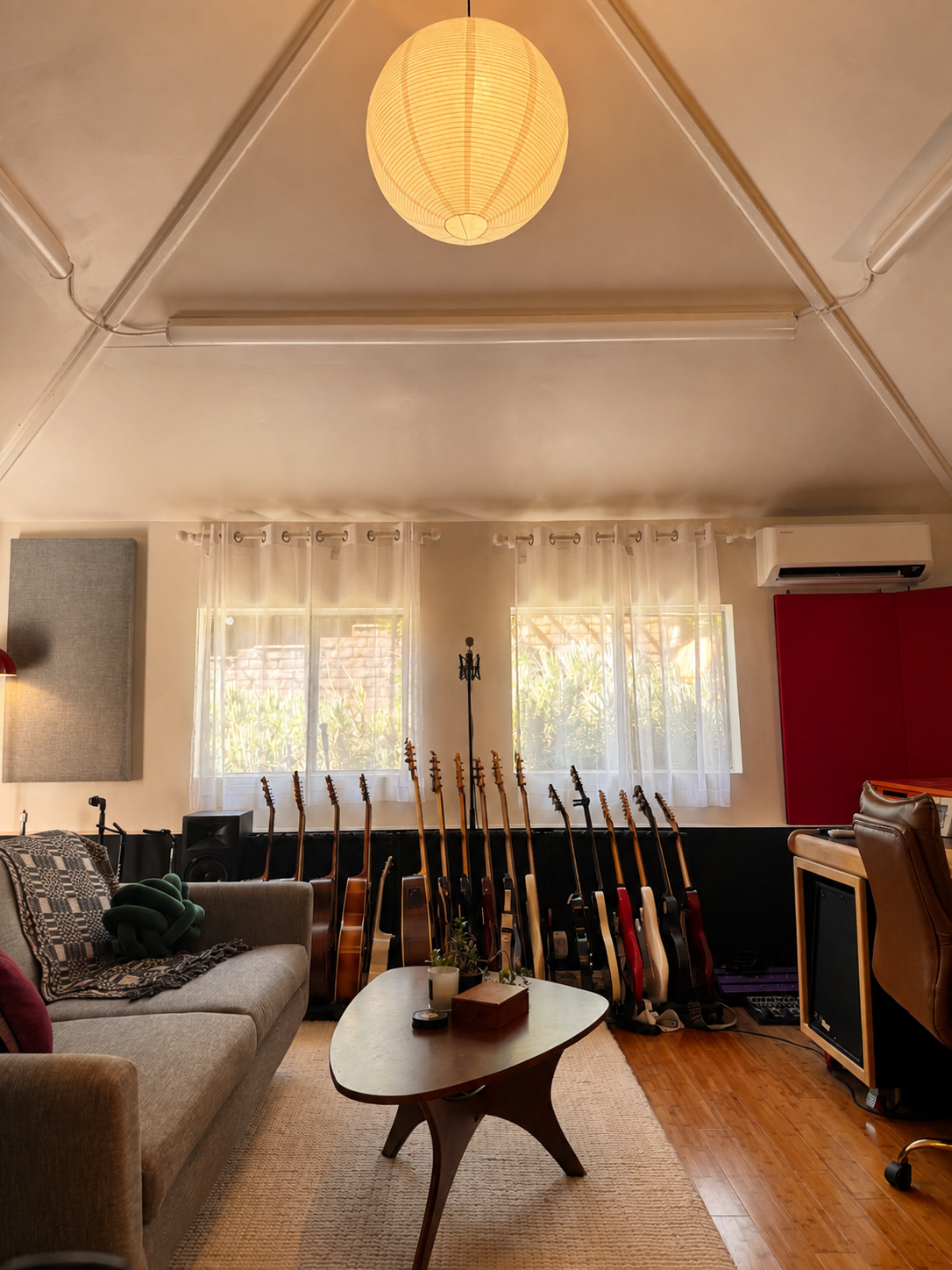 A music studio with several guitars lined up against a wall in front of two windows with sheer curtains allowing natural light, a beige sofa with blankets and pillows, a mid-century coffee table with plants and a box, and recording equipment, with a ceiling lamp hanging from the sloped ceiling.