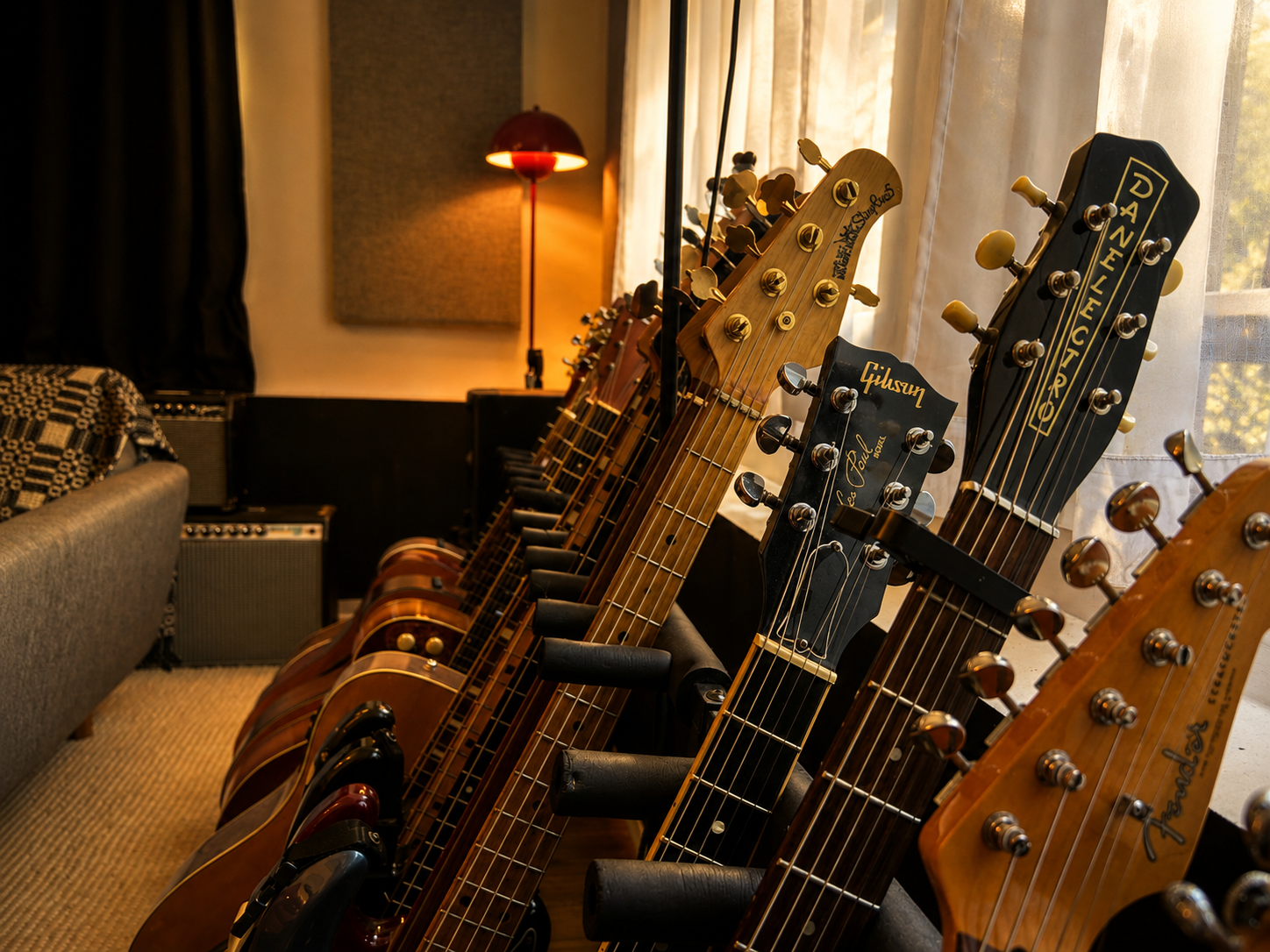 A row of guitar headstocks, including Gibson and Fender, facing a window with curtains in a cozy room with a lamp and an amplifier.