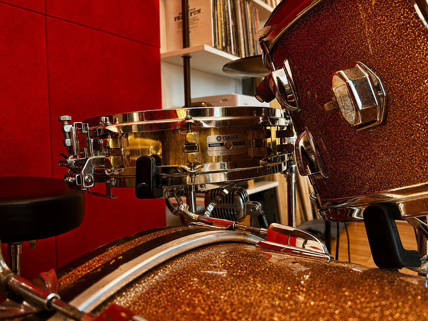 Close-up of a vintage Yamaha drum set with glittery brown finish and chrome hardware, showing drums and part of a drum throne in a room with red and white walls and a bookshelf in the background.