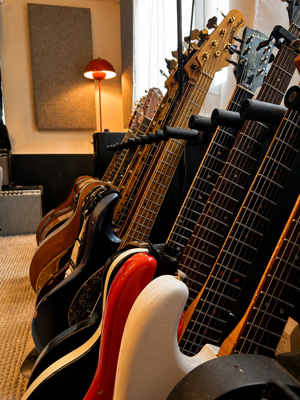 A row of electric guitars and acoustic guitars on stands in a music studio, with a wall lamp and soundproofing panel in the background.