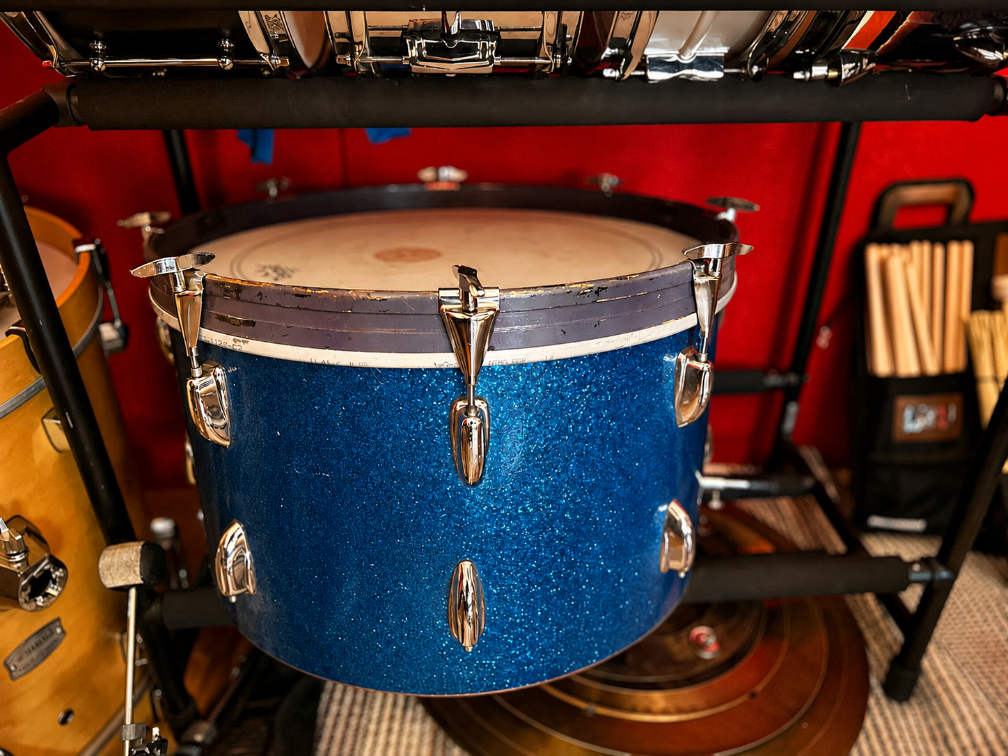 A blue sparkly drum with metal hardware mounted on a black drum rack, with other drums and percussion instruments visible in the background.