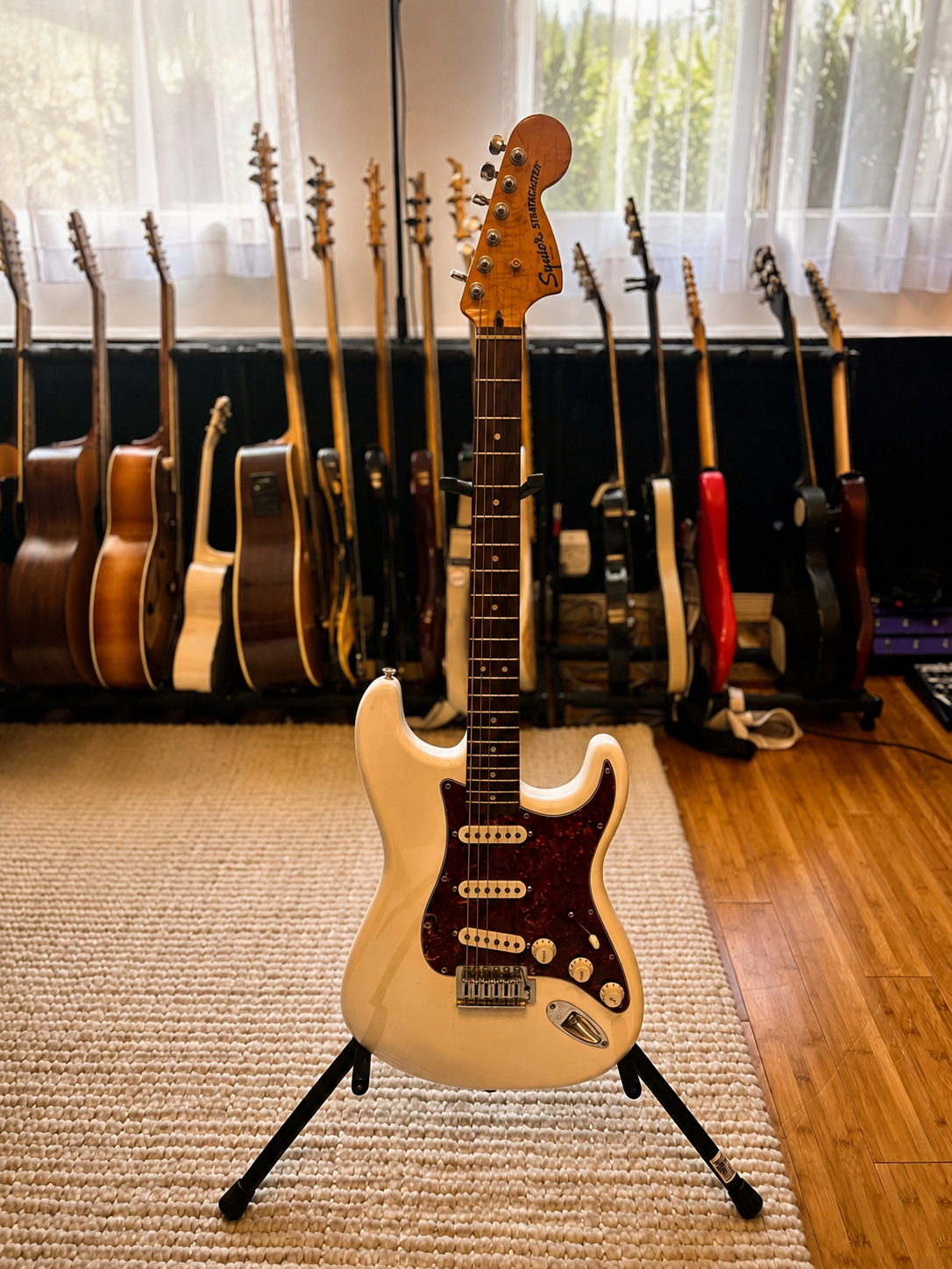 A white vintage-style electric guitar on a stand in a music room, with multiple guitars hanging on a rack behind it and a window with white curtains.