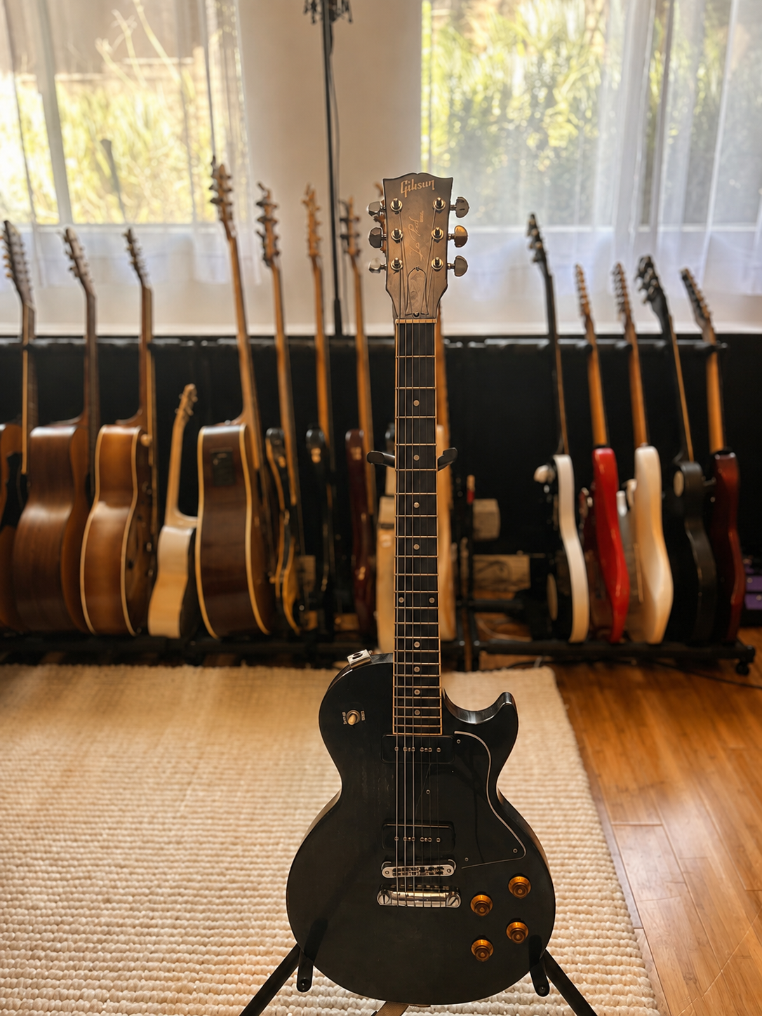 A black Gibson electric guitar on a stand in front of a rack of acoustic and electric guitars in a room with large windows.