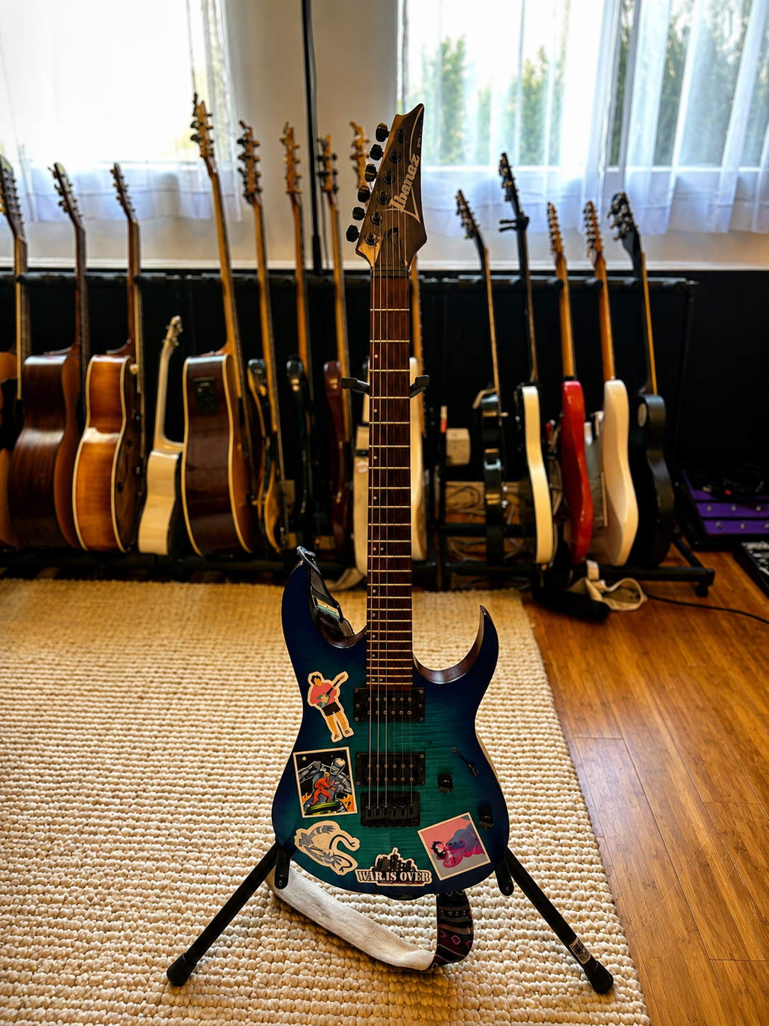An electric guitar with stickers, standing on a guitar stand on a woven mat. In the background, multiple acoustic and electric guitars are hanging on the wall near a window with white curtains.