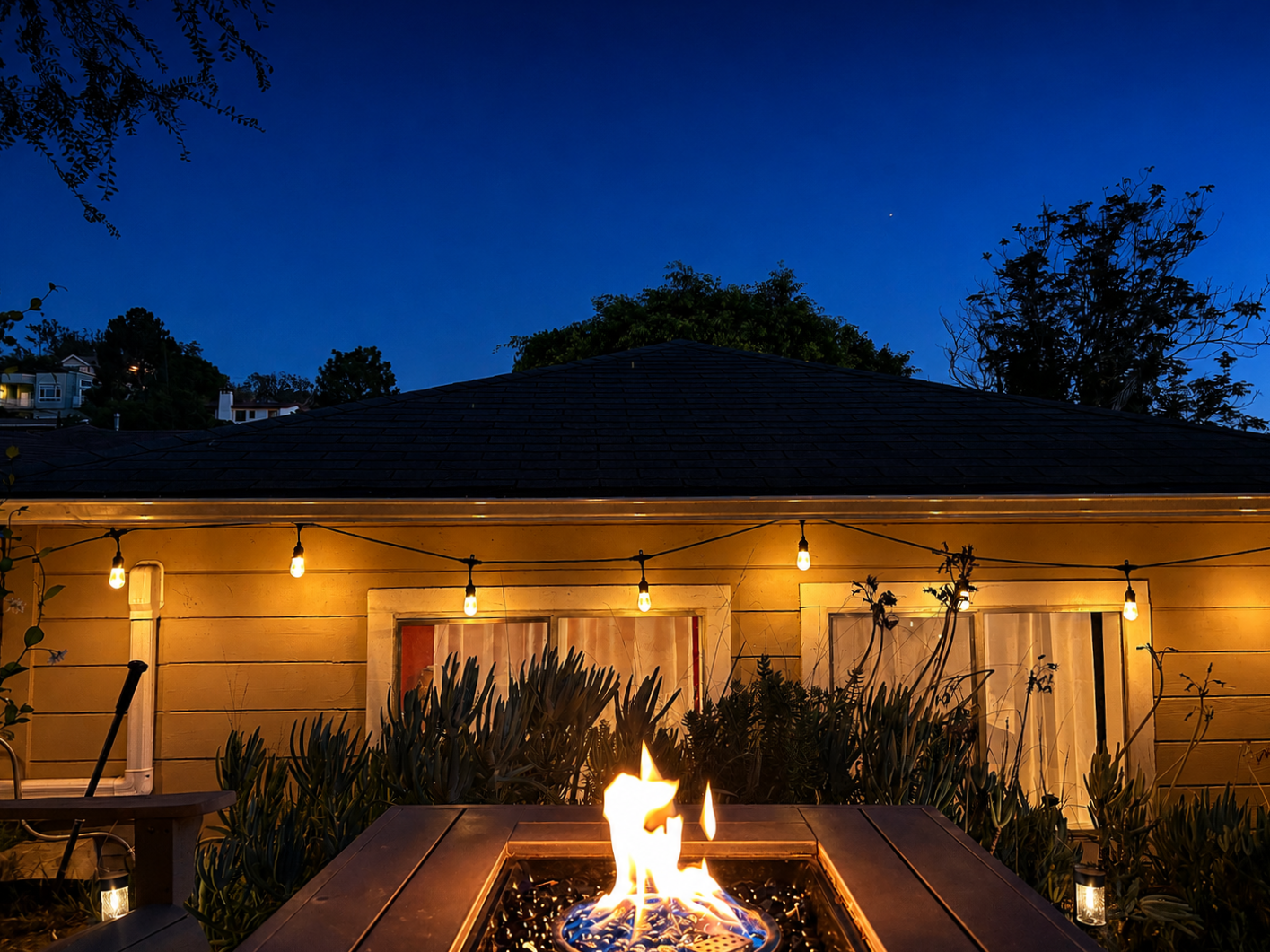 Nighttime backyard scene with a fire pit in the foreground and a house with string lights on the patio. The sky is dark blue with a few stars visible, and trees are silhouetted in the background.
