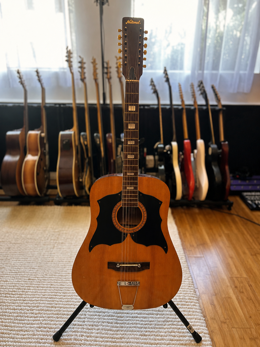 An acoustic guitar with a black pickguard and a sunburst finish, standing on a stand in a music room with a row of electric and acoustic guitars in the background.