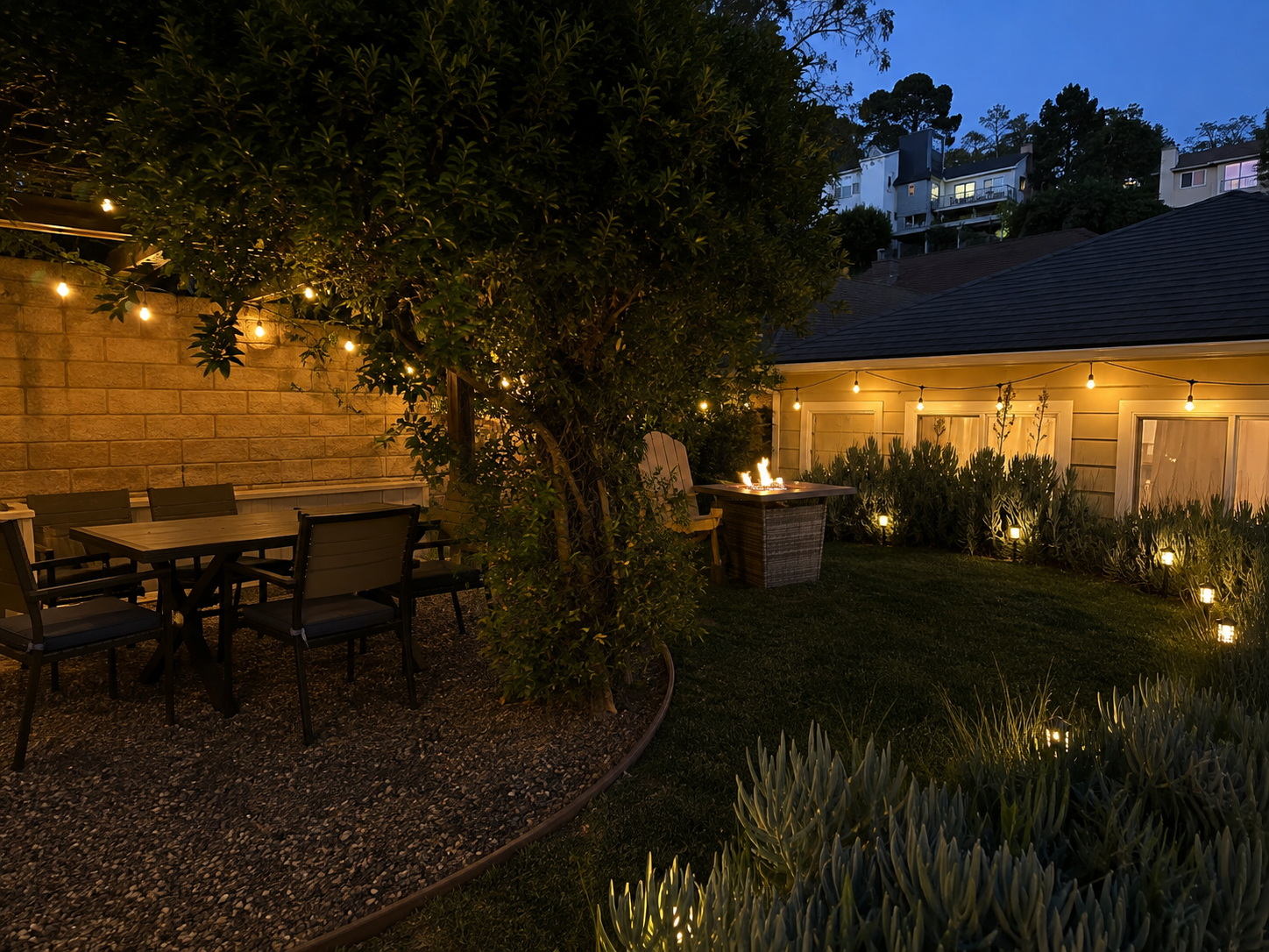 A backyard patio at dusk with string lights above a dining table and chairs, a fire pit near a house, and garden plants along the perimeter.