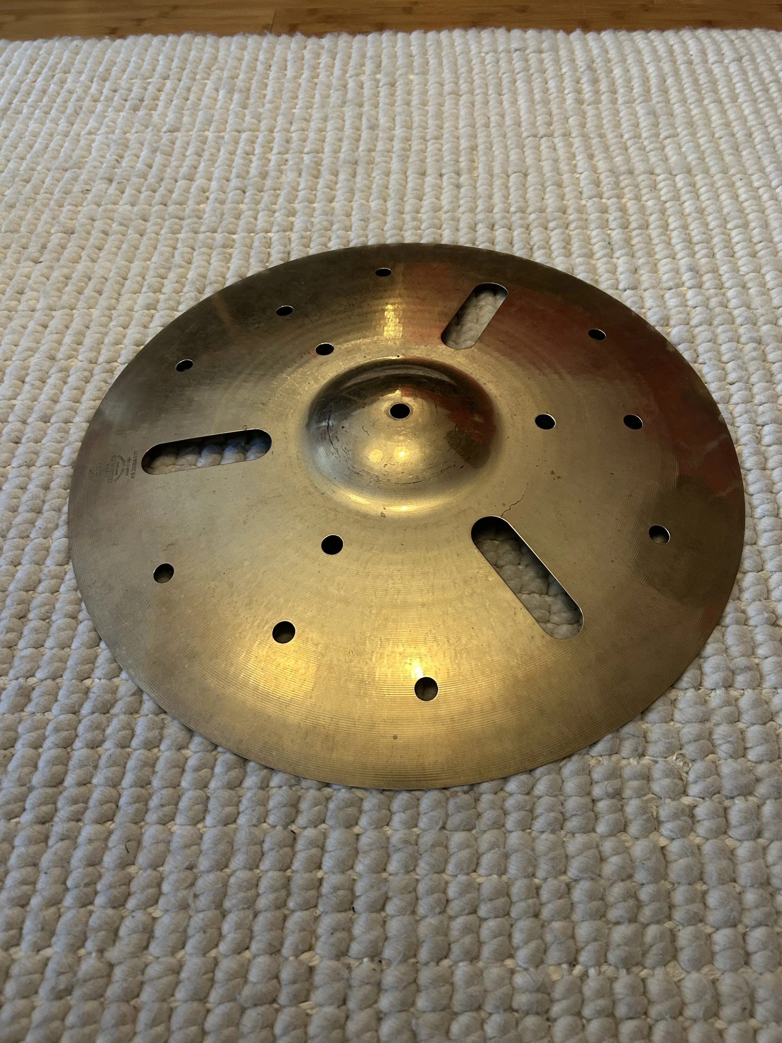 A round metal object with multiple holes and slots, resting on a textured white rug