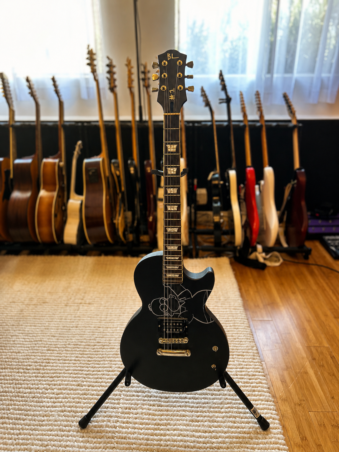 Black electric guitar with cartoon character drawing on the body, standing on a tripod in a music room, with multiple acoustic and electric guitars on stands in the background near a window.