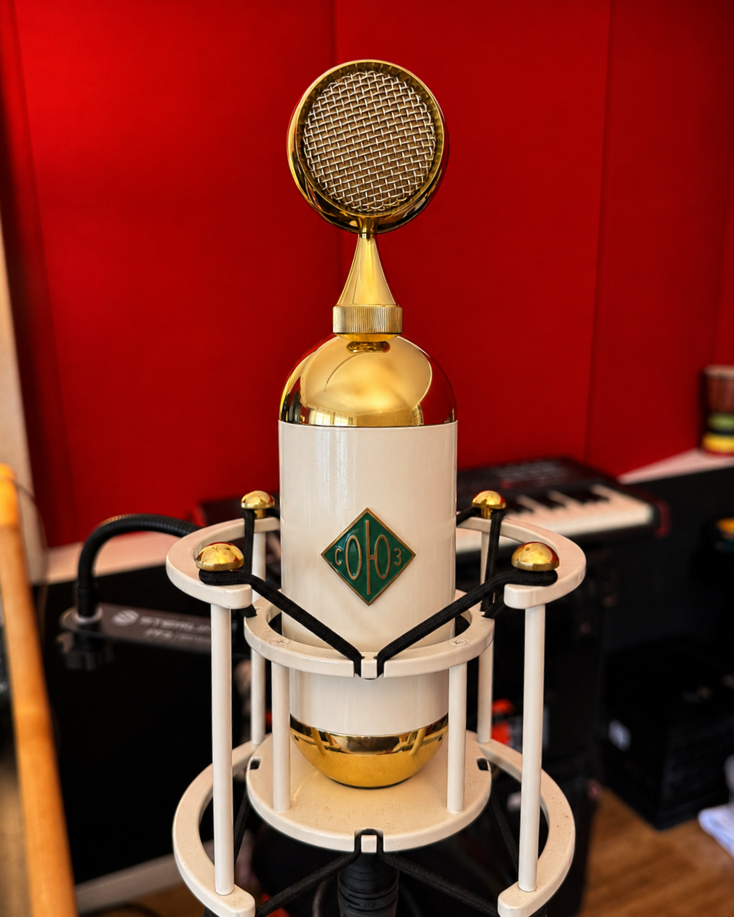A vintage large tube microphone with gold accents, surrounded by a circular stand with white and black supports, placed in a recording studio with a red wall.