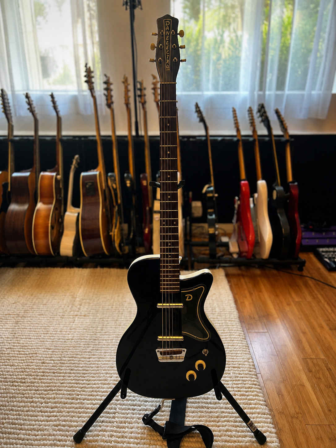 A black electric guitar with a unique shape on a guitar stand in front of a collection of wooden and electric guitars on a rack near a window with curtains.