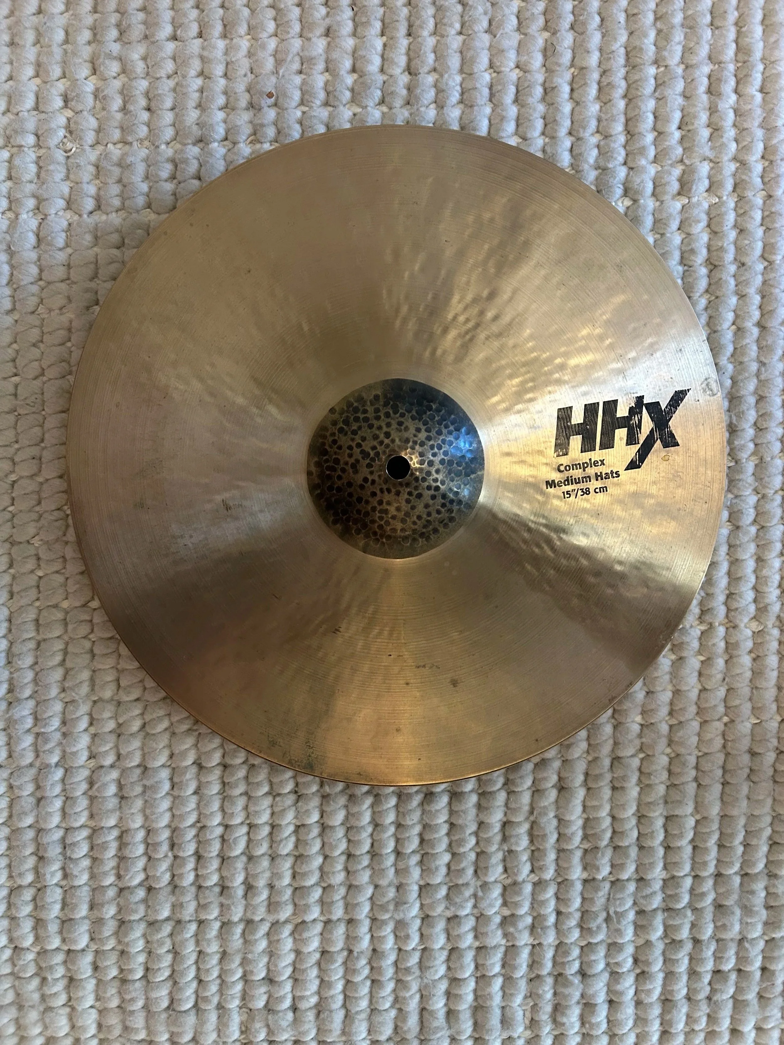 A brass cymbal with the brand name 'HI-HAT' and size 15 inches (38 cm), resting on a textured beige carpet.