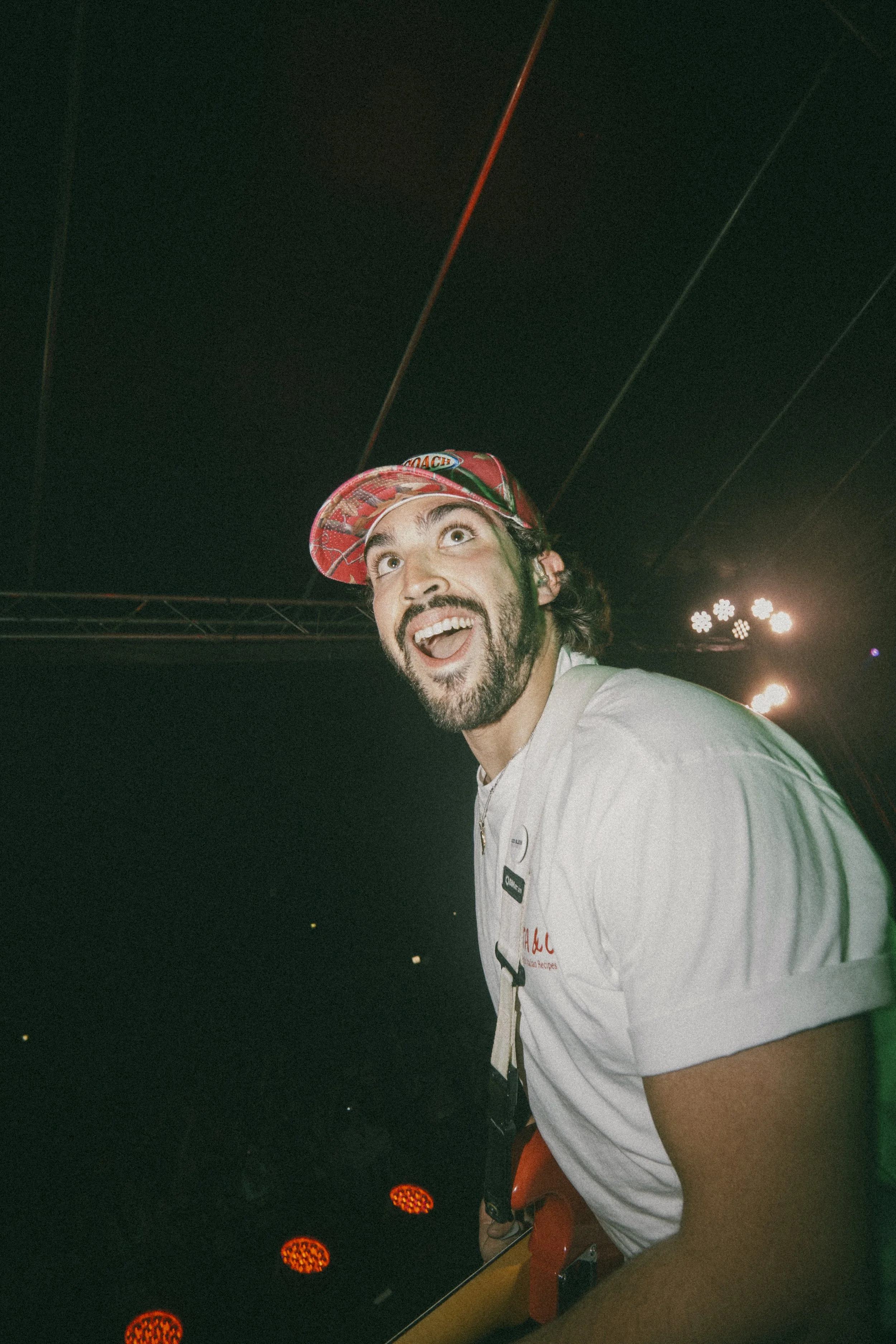A man with a beard and long hair wearing a red cap, white shirt, and orange headphones around his neck, smiling and looking upward while sitting at an arcade game with orange lights.