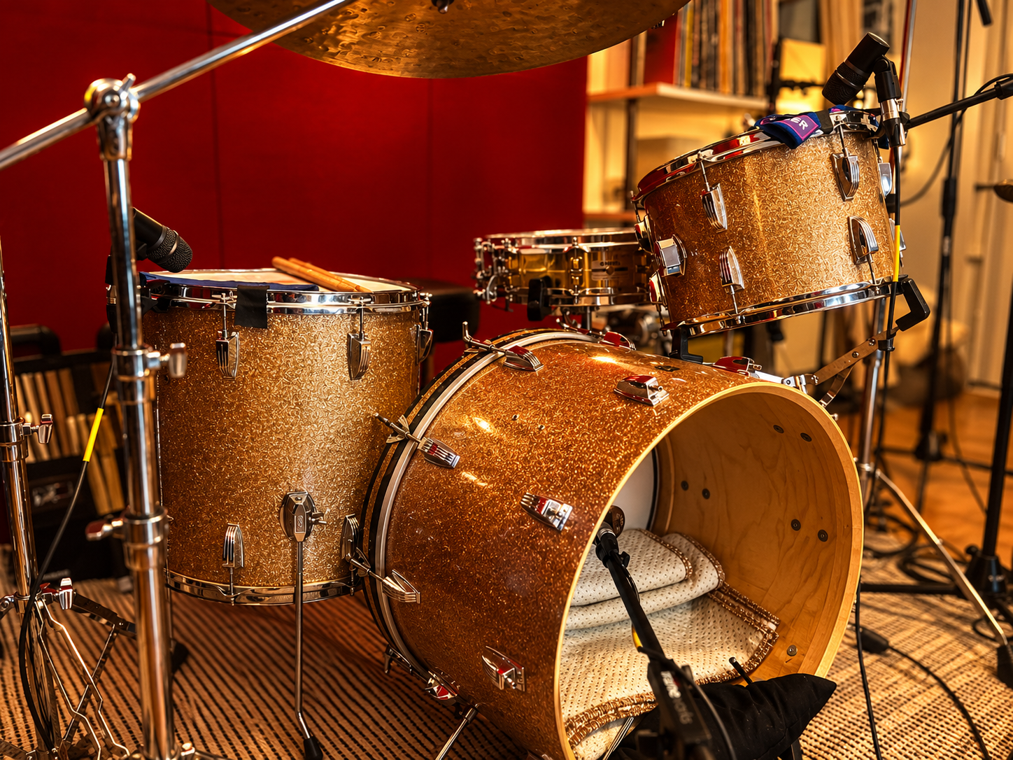 A set of gold glittering drum kit, including a bass drum, two tom-toms, and various cymbals, with microphones and stands in a music studio or rehearsal space.