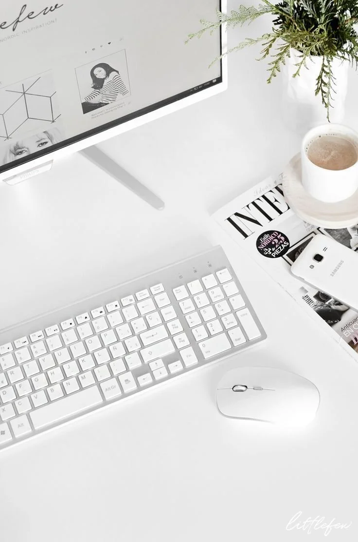 A white desk setup with a computer monitor, keyboard, mouse, a cup of coffee, a magazine, a smartphone, and a potted plant.