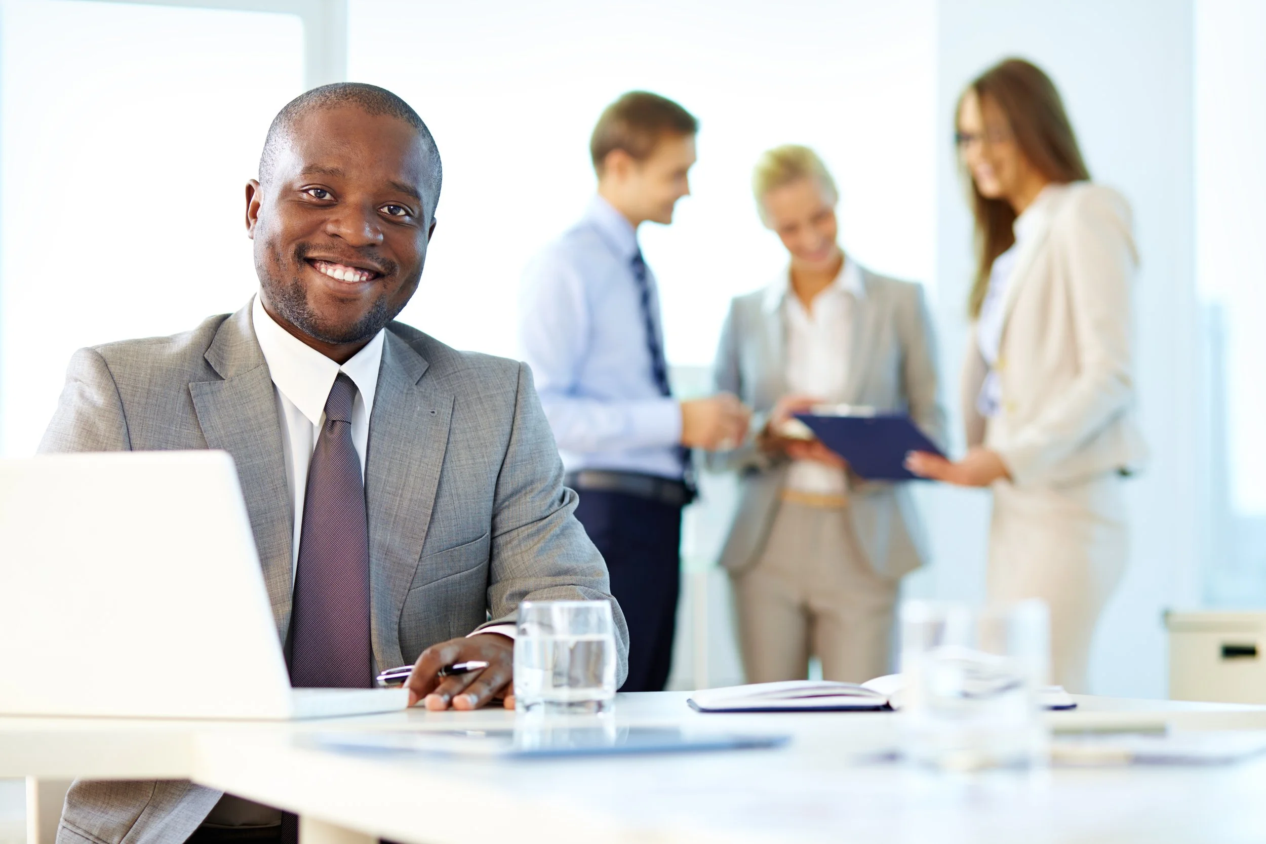 A smiling African American man in a light gray suit and tie sitting at a table with a glass of water, with a group of four professionally dressed people in the background engaged in conversation in a bright, modern office setting.