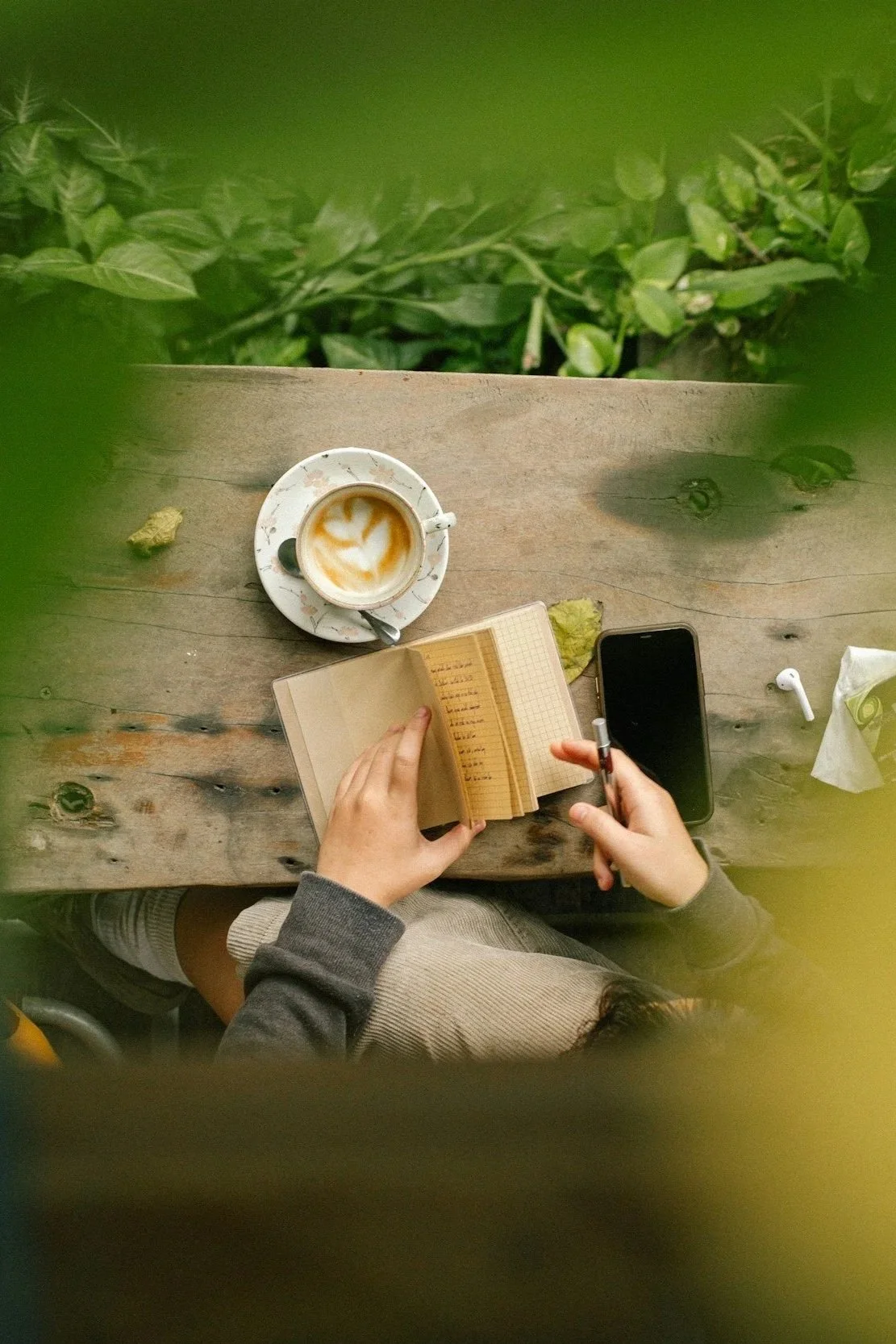 Overhead view of a woman at a wooden table with a cup of coffee, an open notebook, a smartphone, and a pair of AirPods. Green foliage frames the scene at the top.