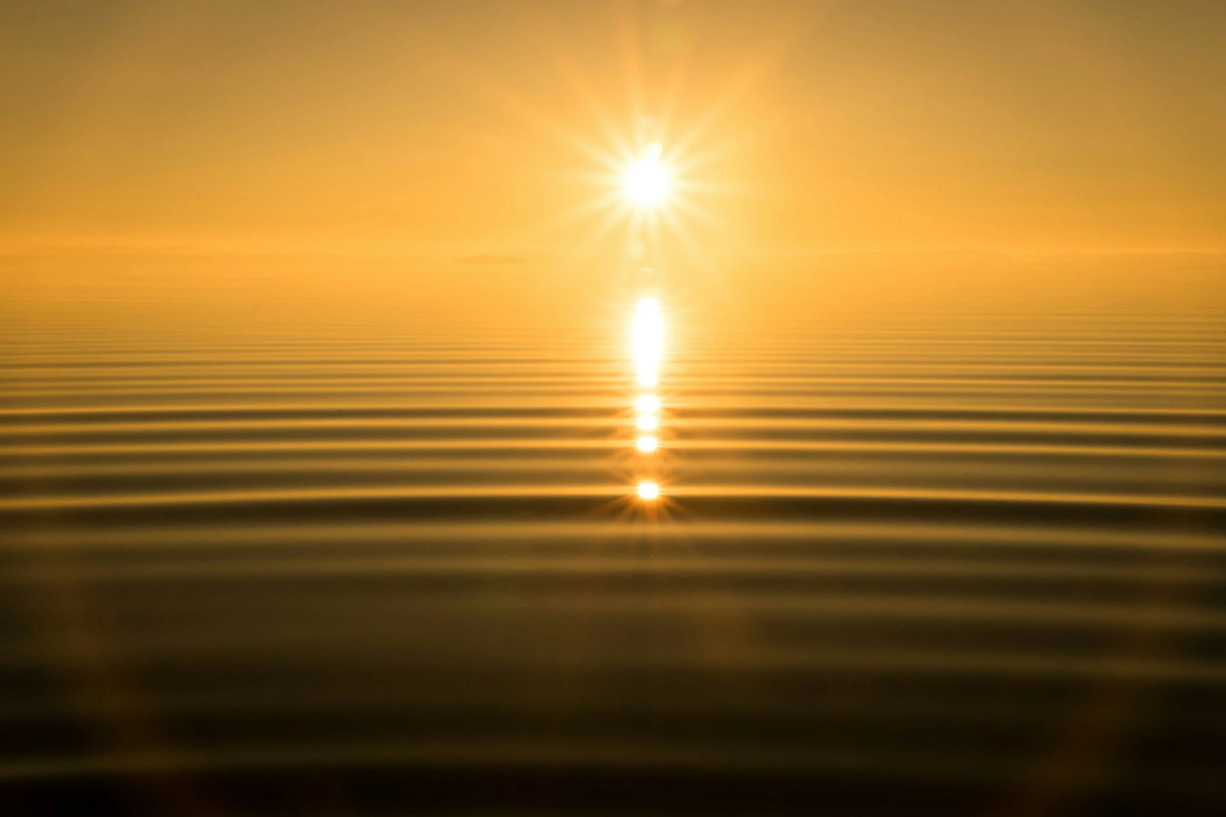 A sunset or sunrise over a calm body of water with the sun's reflection visible on the surface, creating a golden glow and gentle ripples.