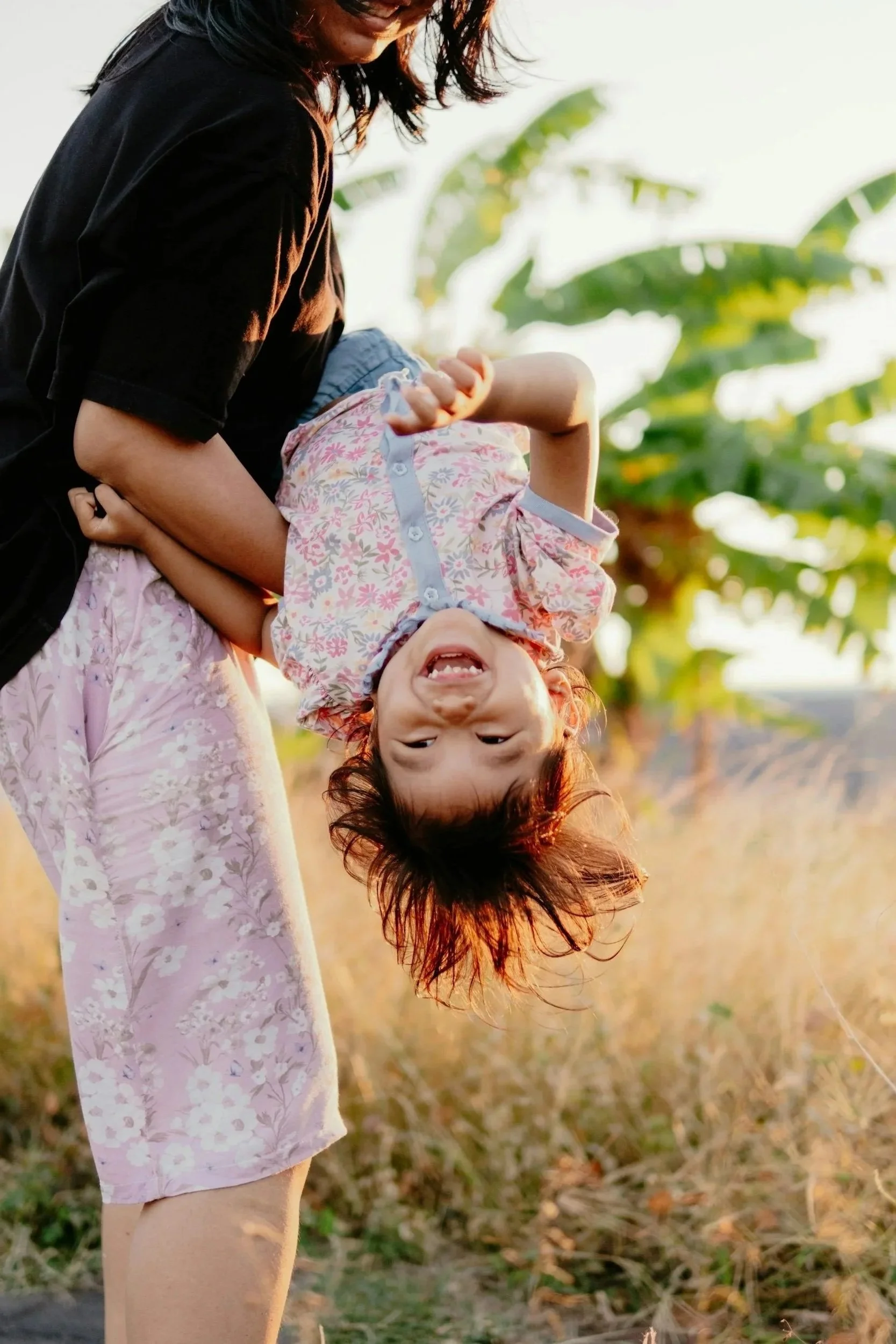A woman in a black top holding a smiling girl upside down outdoors in a field during sunset.