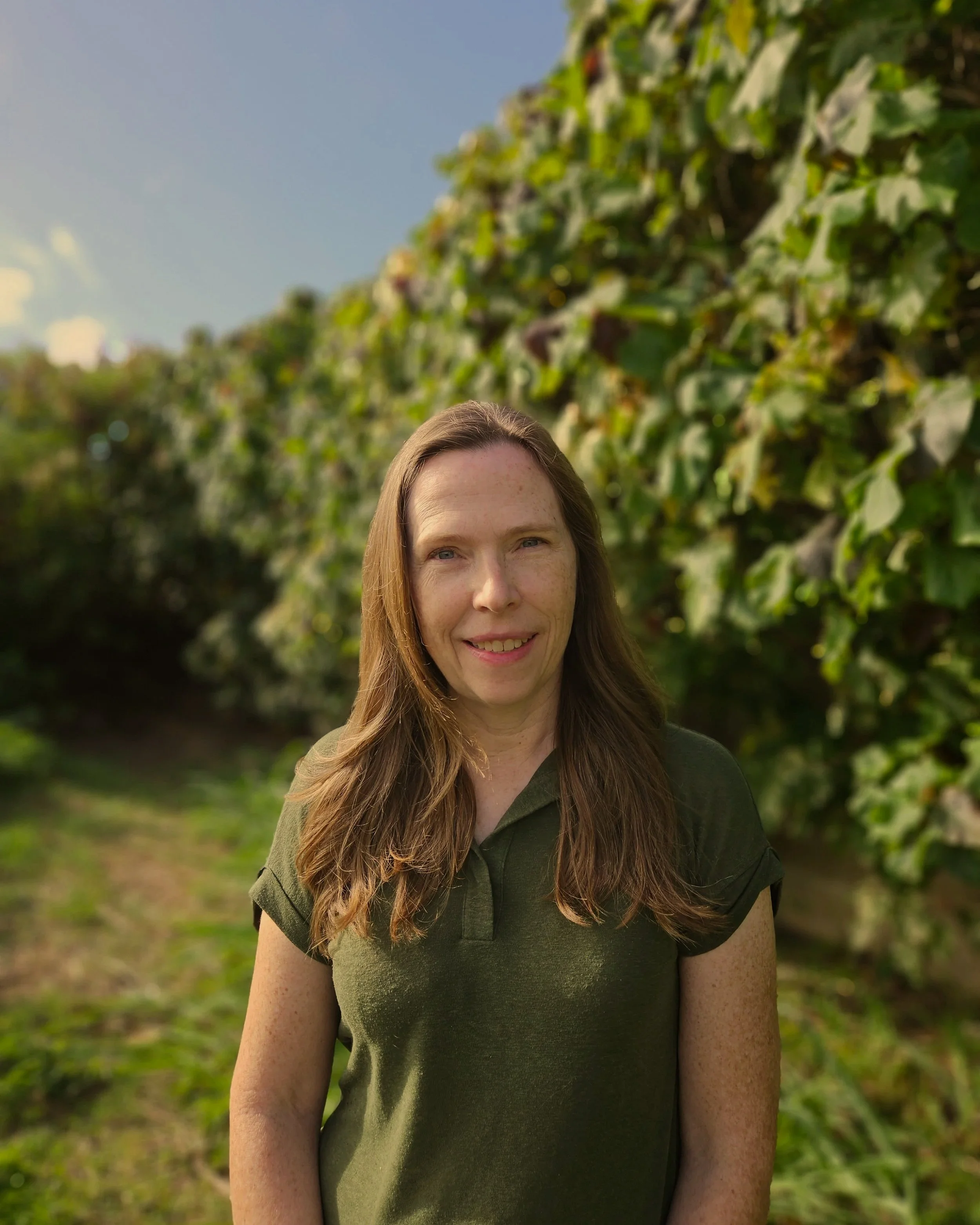 A woman with long brown hair and light skin, smiling outdoors with green foliage and a clear sky in the background.