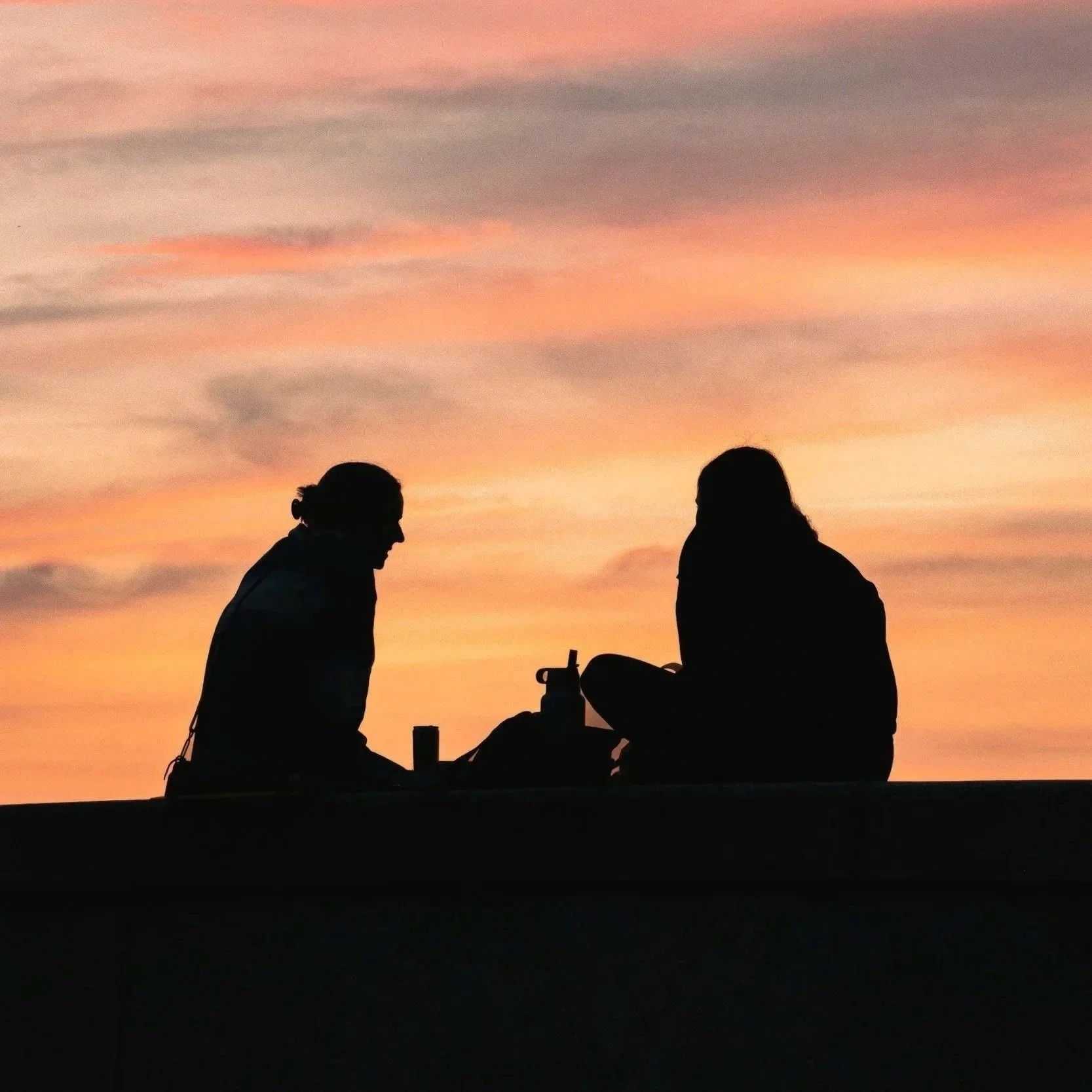 Silhouettes of two women sitting at a table during sunset, with colorful pink and orange clouds in the sky.