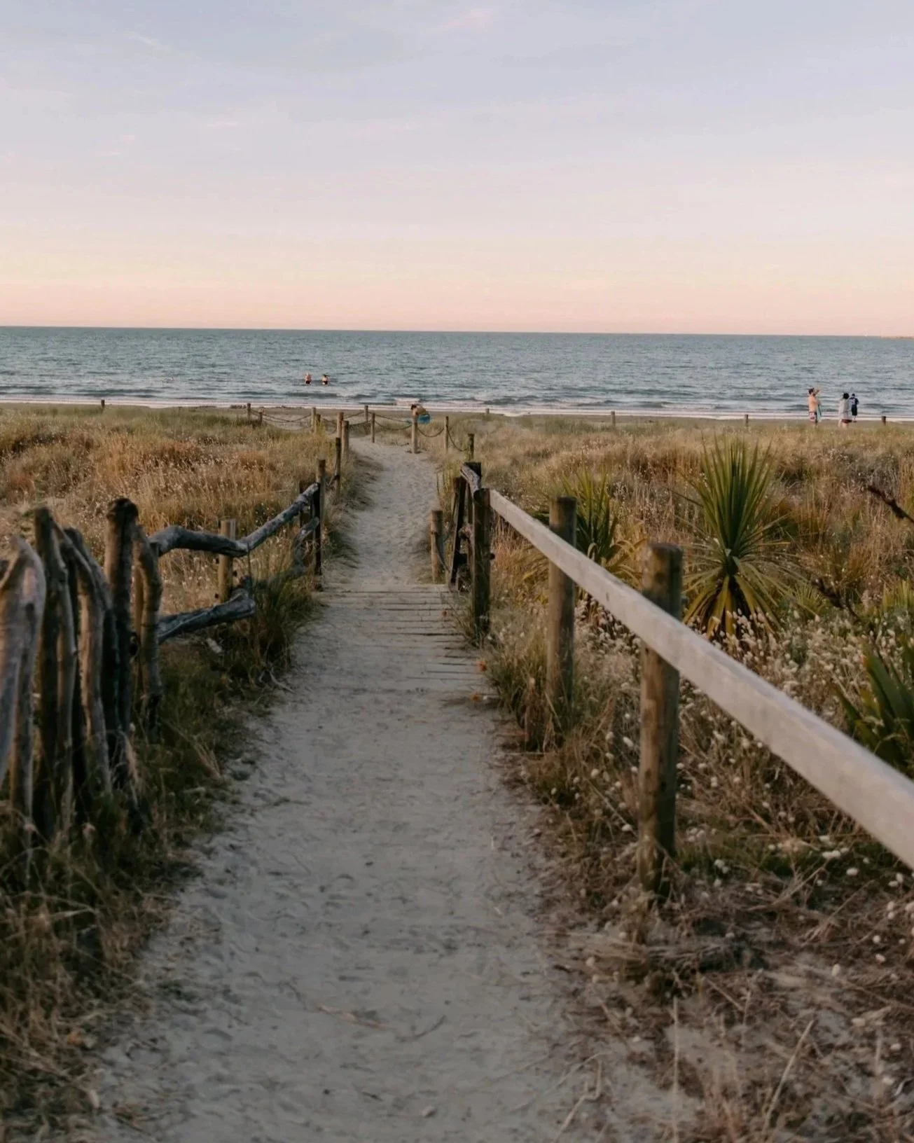A sandy pathway bordered by wooden and rope fencing leads to a beach with grassy dunes and the ocean in the background. People are walking along the shoreline, and some are swimming in the ocean. The sky is mostly clear with some soft clouds, suggesting late afternoon or early evening.