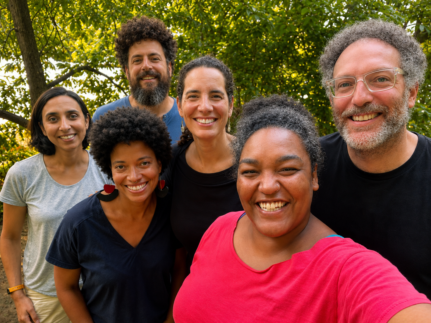 Group of six diverse people smiling outdoors with trees in the background, taking a selfie.