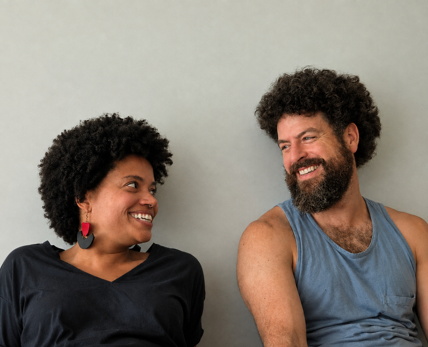 A woman and a man sitting against a plain wall, smiling at each other. The woman has curly black hair, wearing a black shirt and large red and black earrings. The man has curly hair and a beard, wearing a sleeveless blue shirt.