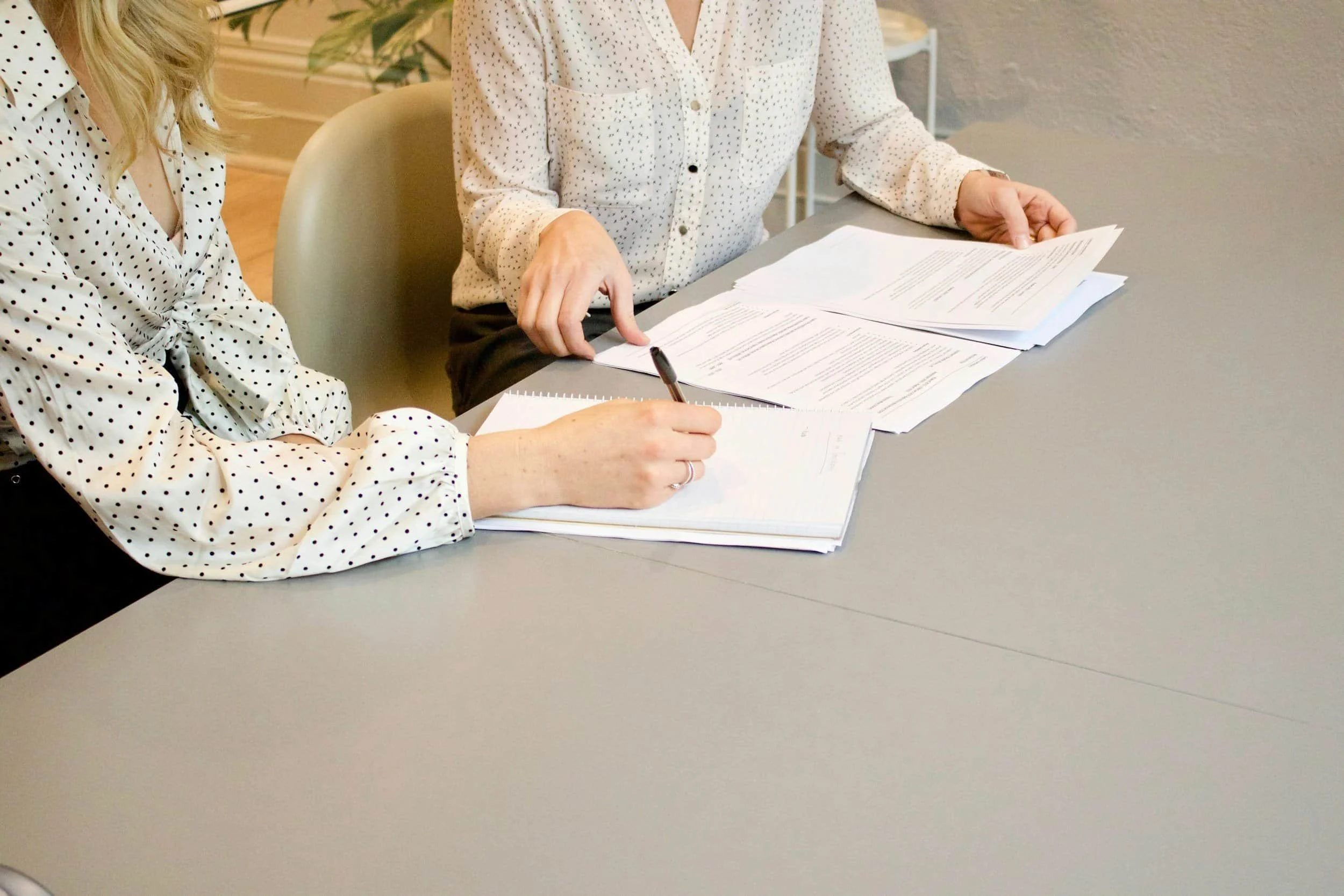 Two women in white blouses with black polka dots working with papers and a pen at a table.