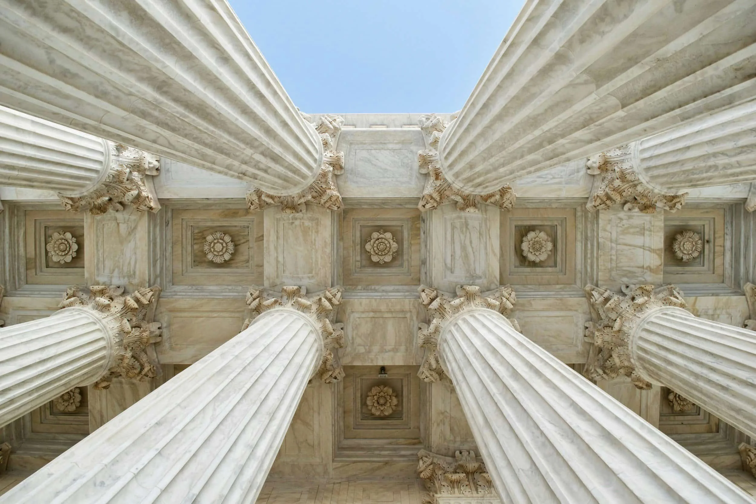 From below view of ancient Greek or Roman marble architecture with tall, fluted columns and ornate capitals, looking up toward the blue sky.