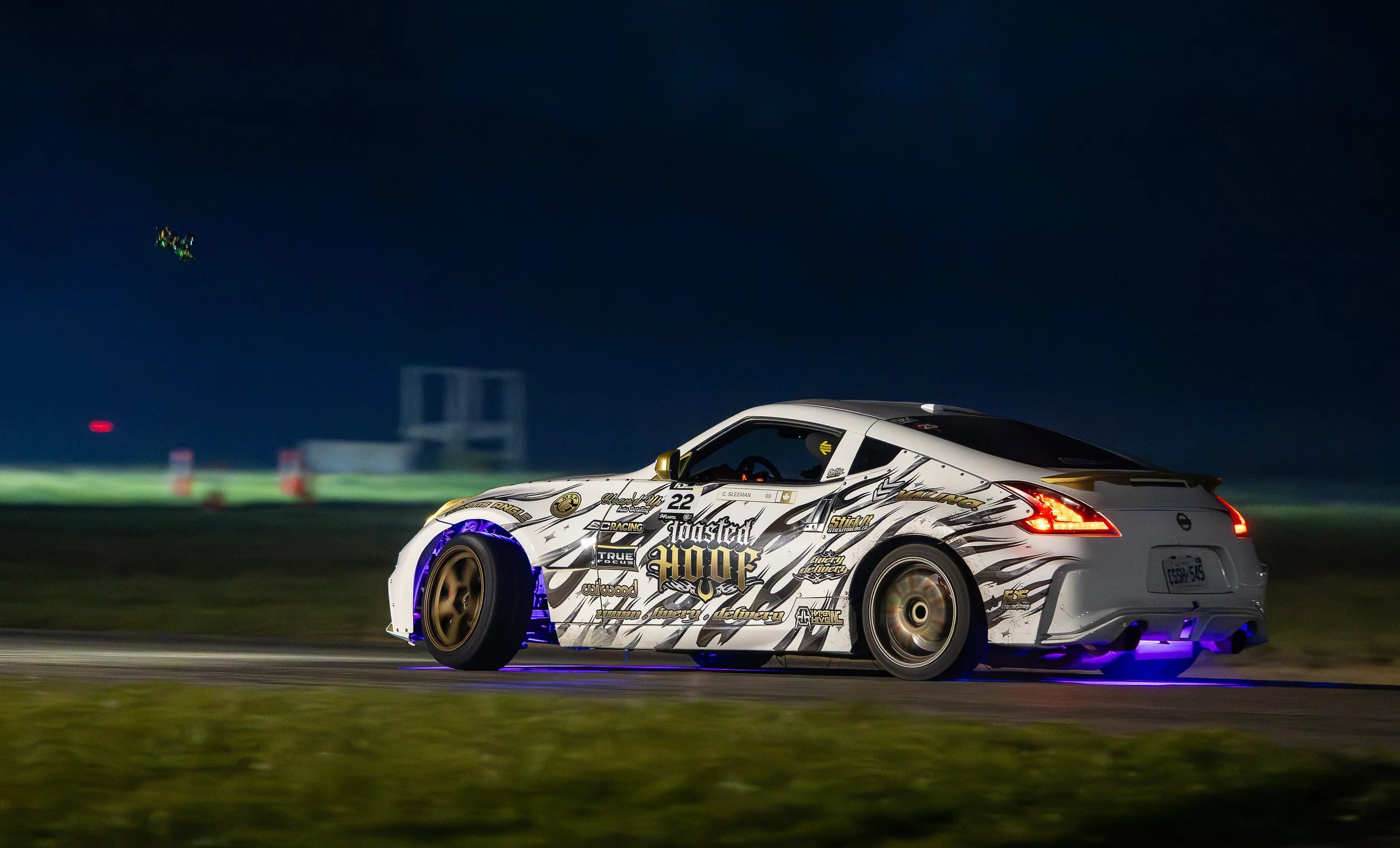 A white race car with black and gold decals and branding, driven by a person in a helmet, racing on a track at night with colorful lights in the sky.