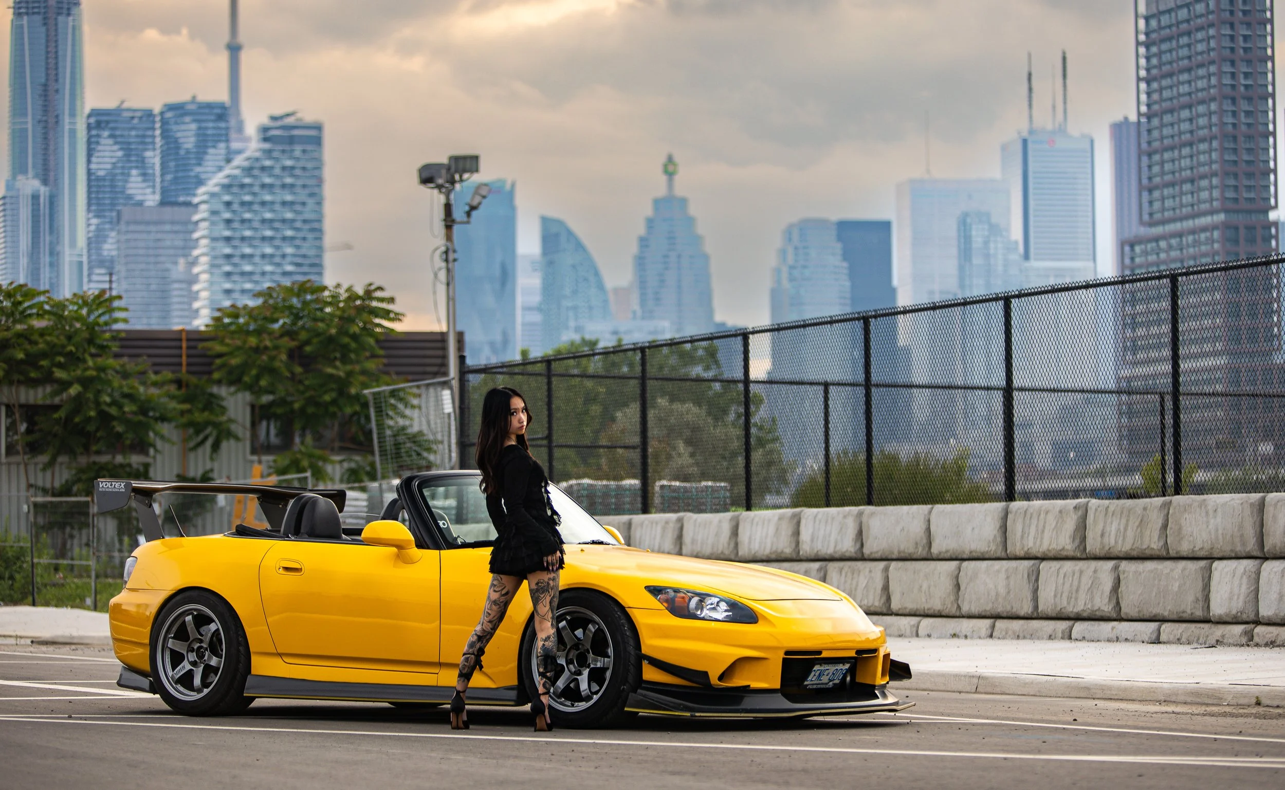 A woman with long dark hair and tattoos on her legs standing next to a yellow sports car in a parking lot with a city skyline in the background.
