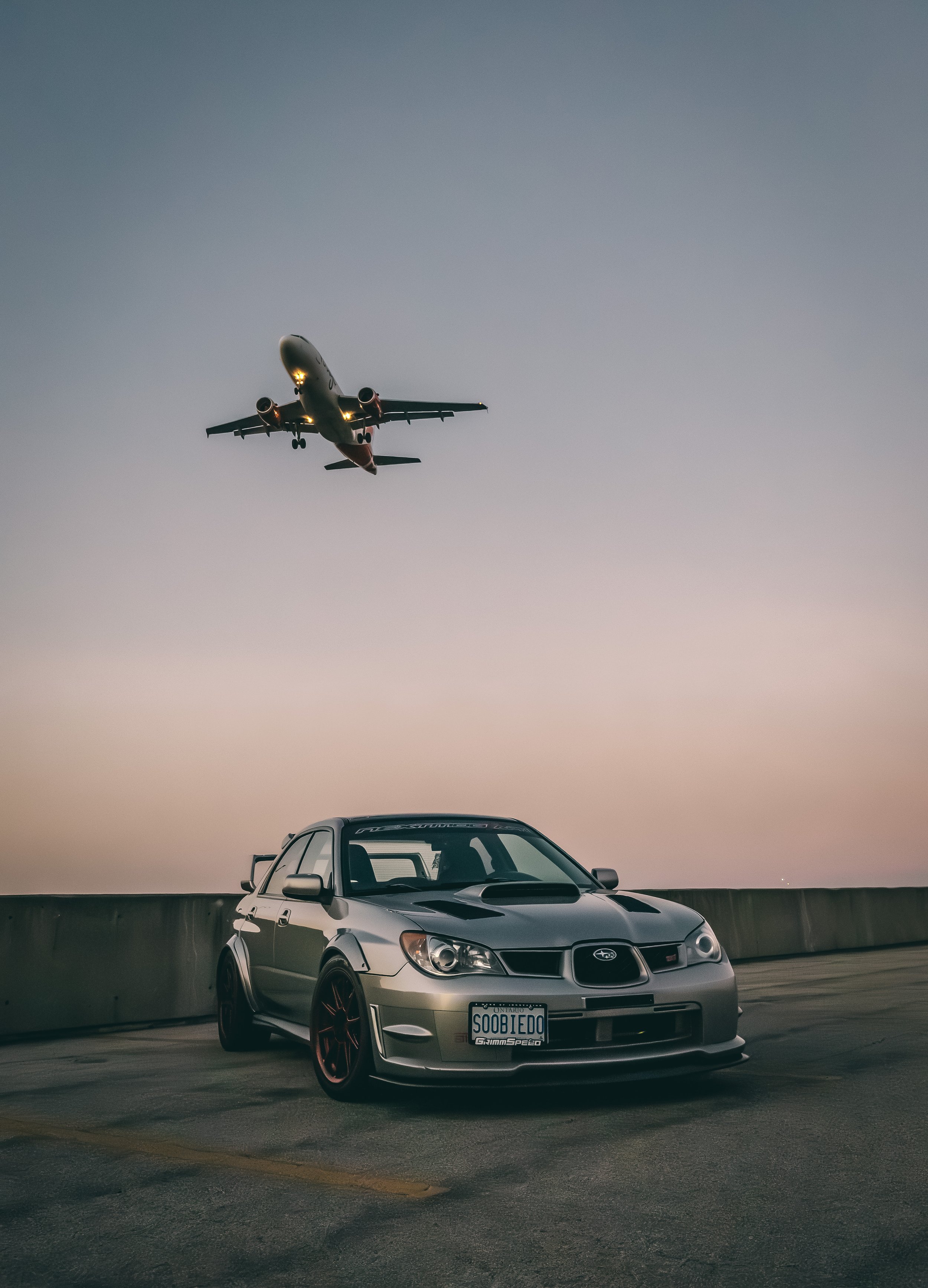 A silver Subaru car with red rims parked on a rooftop parking lot with a concrete barrier, under a gradient sky at dusk. A commercial airplane is flying overhead in the sky.