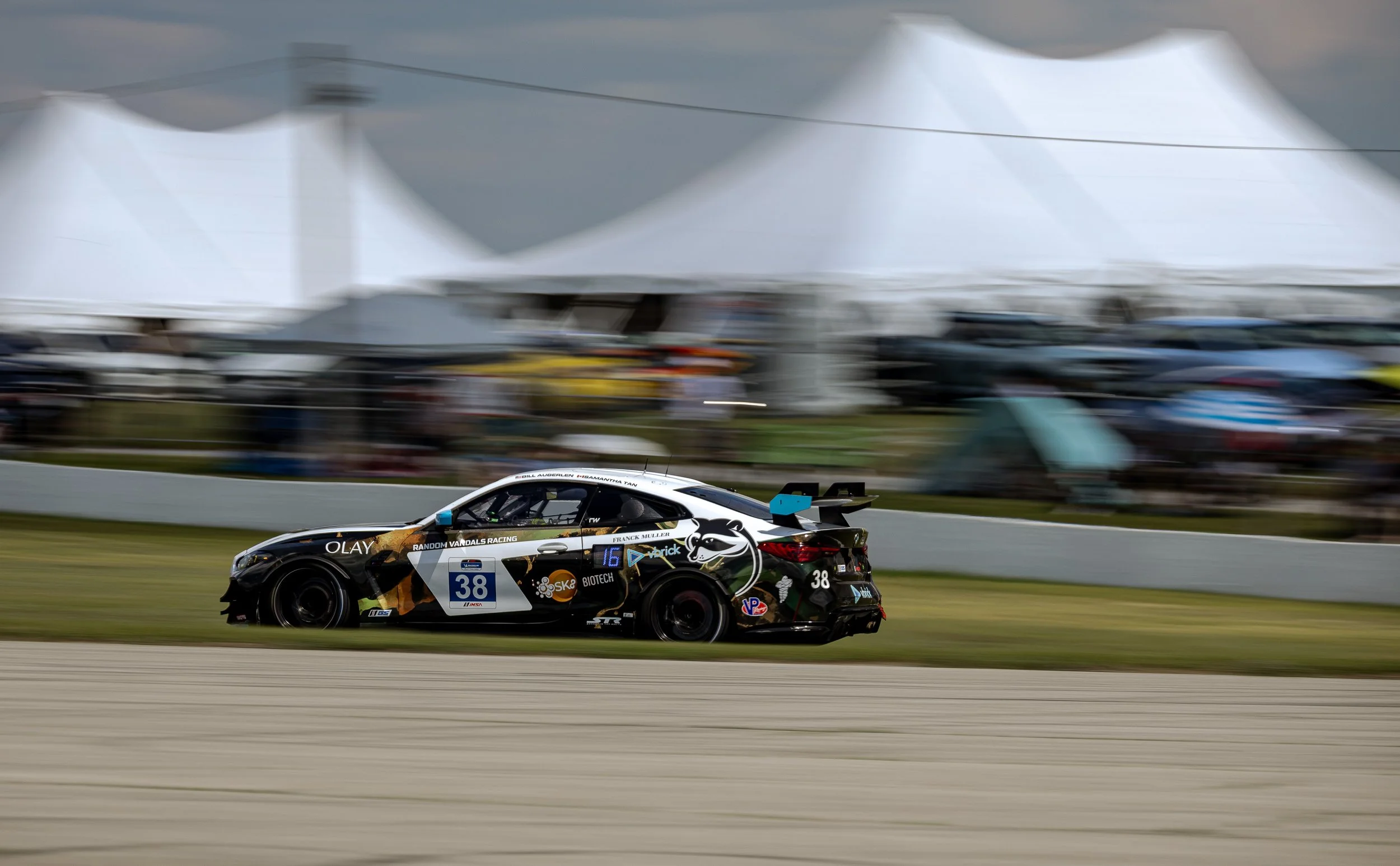 Race car with black, white, and gold livery speeding on a race track with blurred background tents and cars.