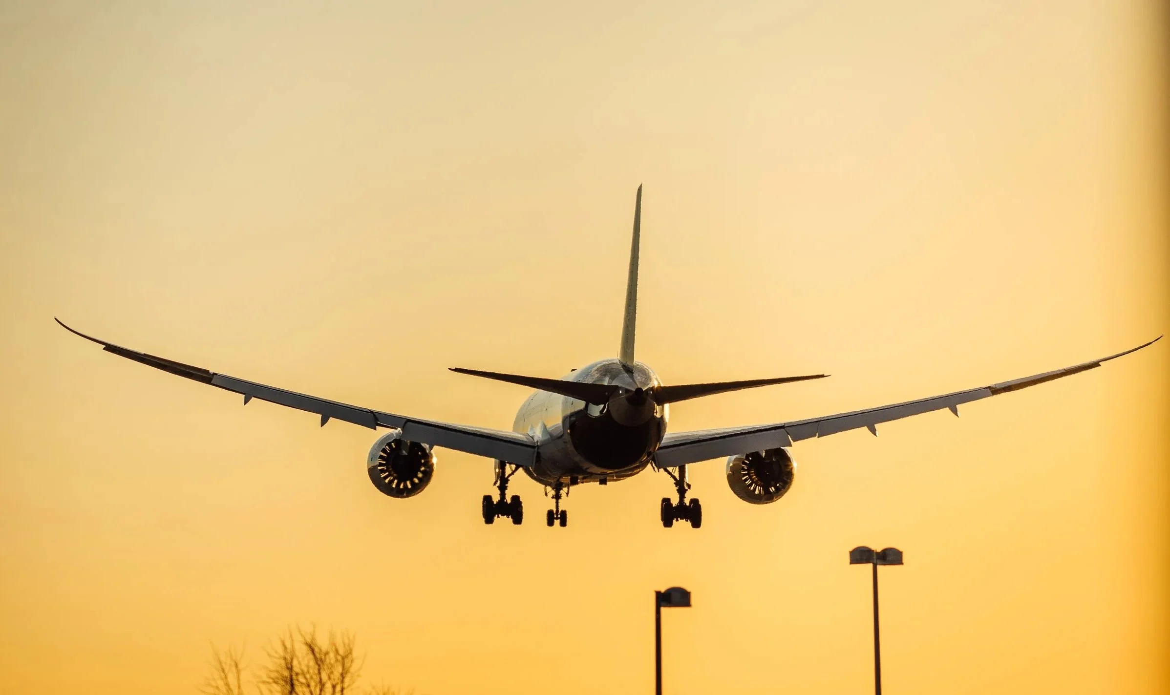 A commercial airplane flying low against a yellowish sky during sunset, with landing gear extended and engines visible beneath the wings.
