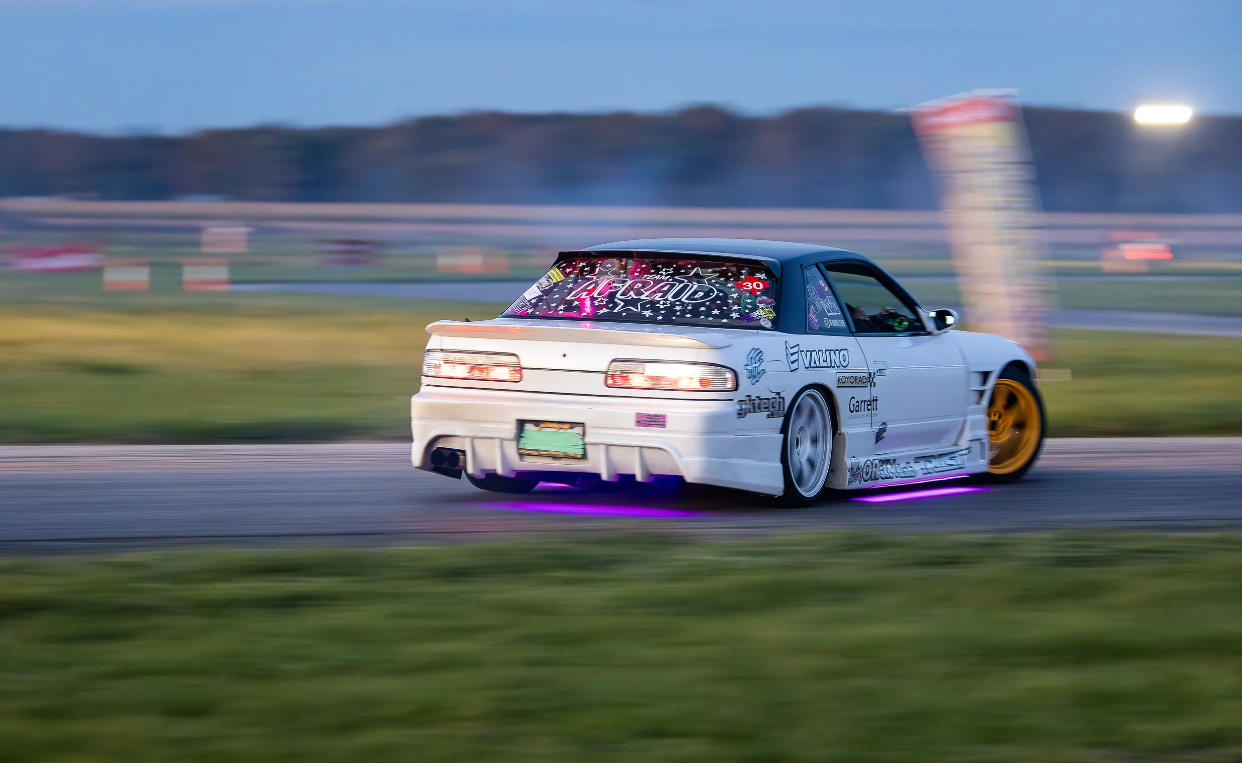 A white racing car with sponsor logos, yellow wheels, and neon underglow lights, racing on a track with a blurred background and motion streaks indicating high speed.