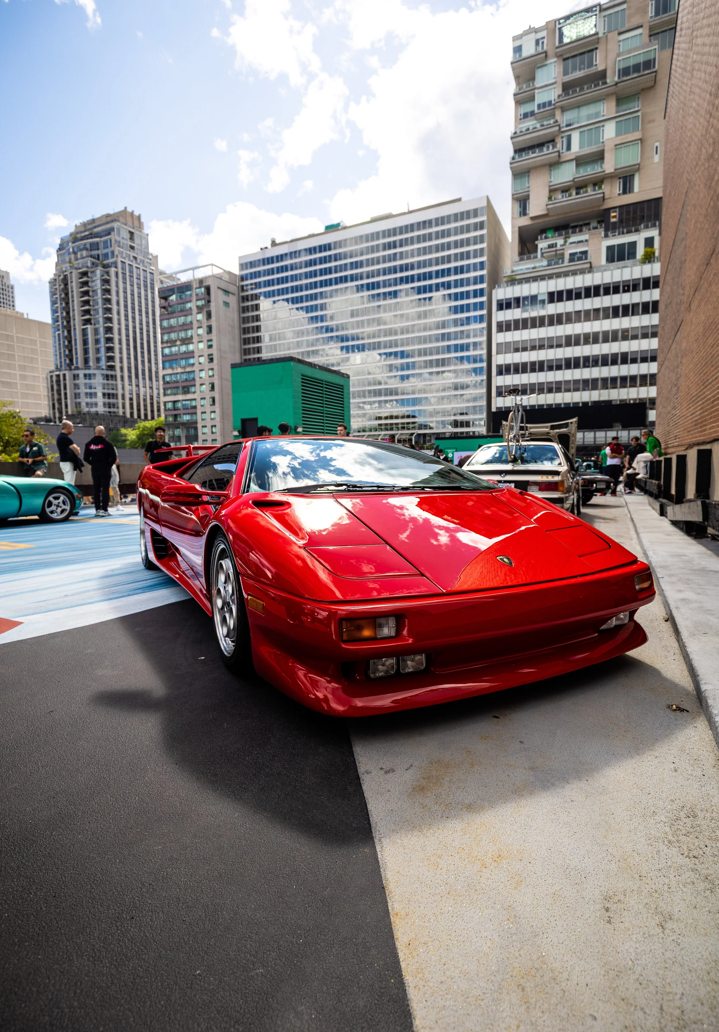A red sports car parked in an urban parking lot with tall modern buildings and people walking around.