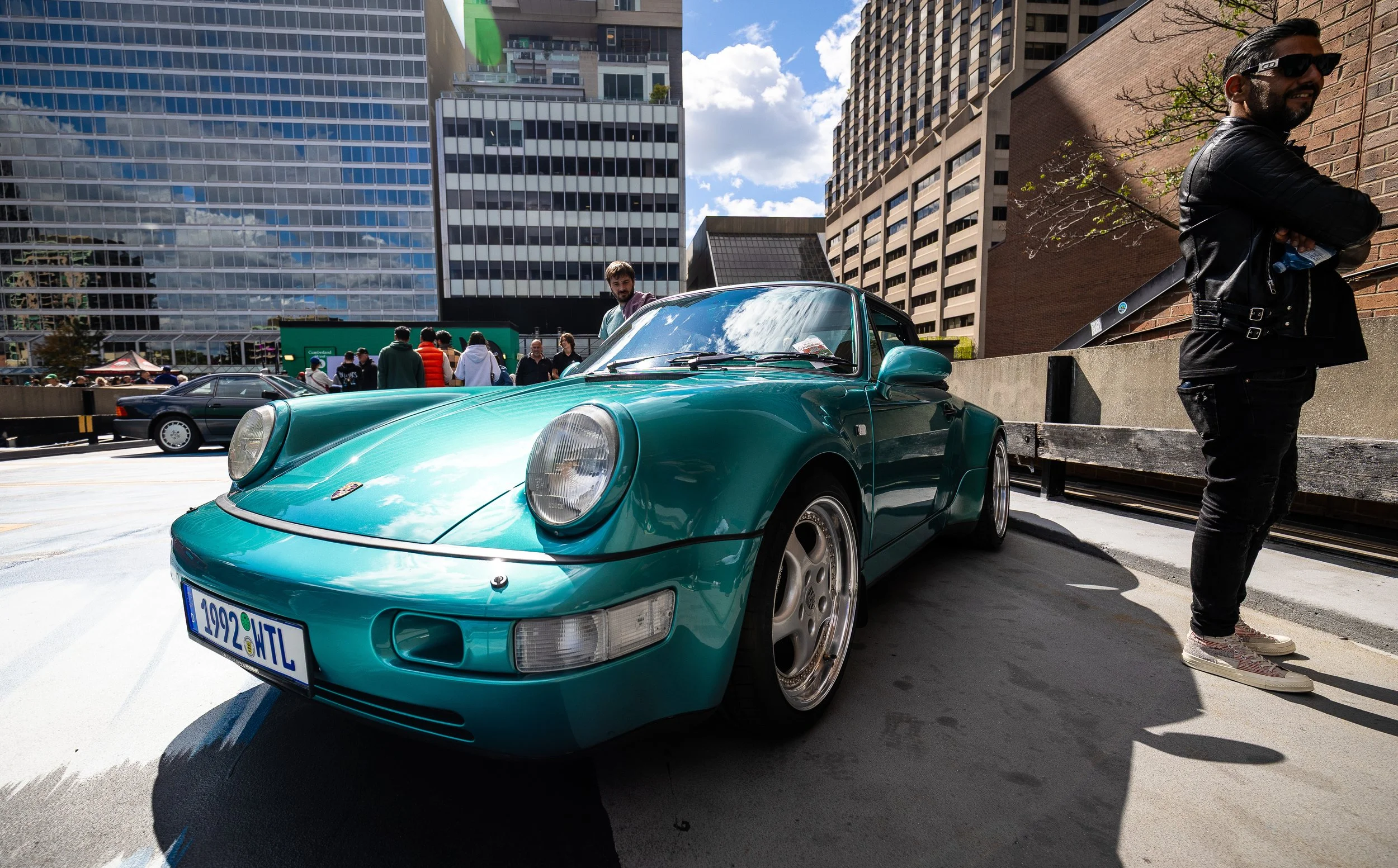 A teal Porsche sports car parked on the street during a car show in an urban area with tall modern buildings. A man wearing sunglasses and a leather jacket stands next to a brick wall, holding a water bottle, and other people are seen walking and loo